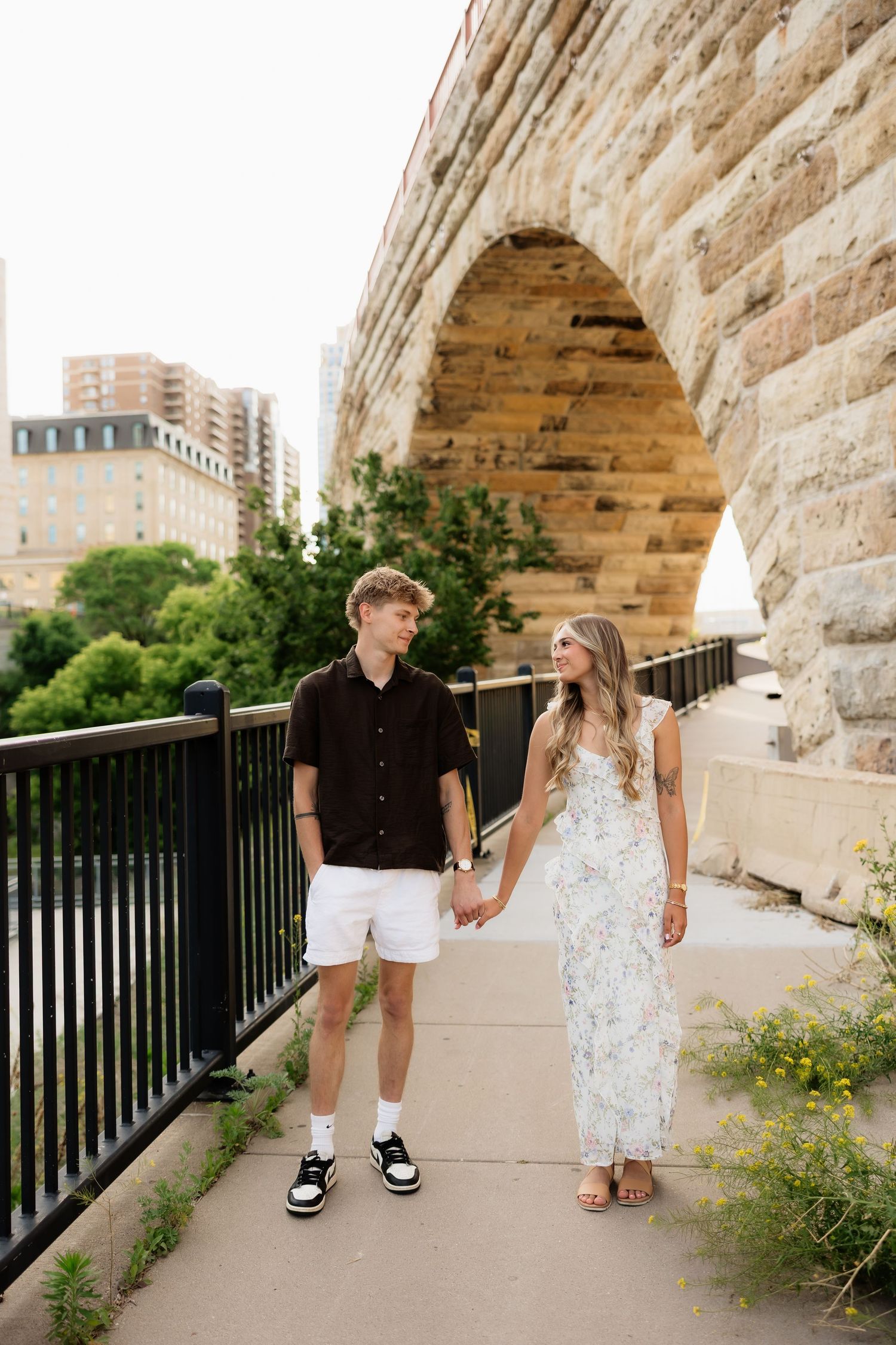 Couple holding hands and walking along a path beside a historic stone arch bridge.
