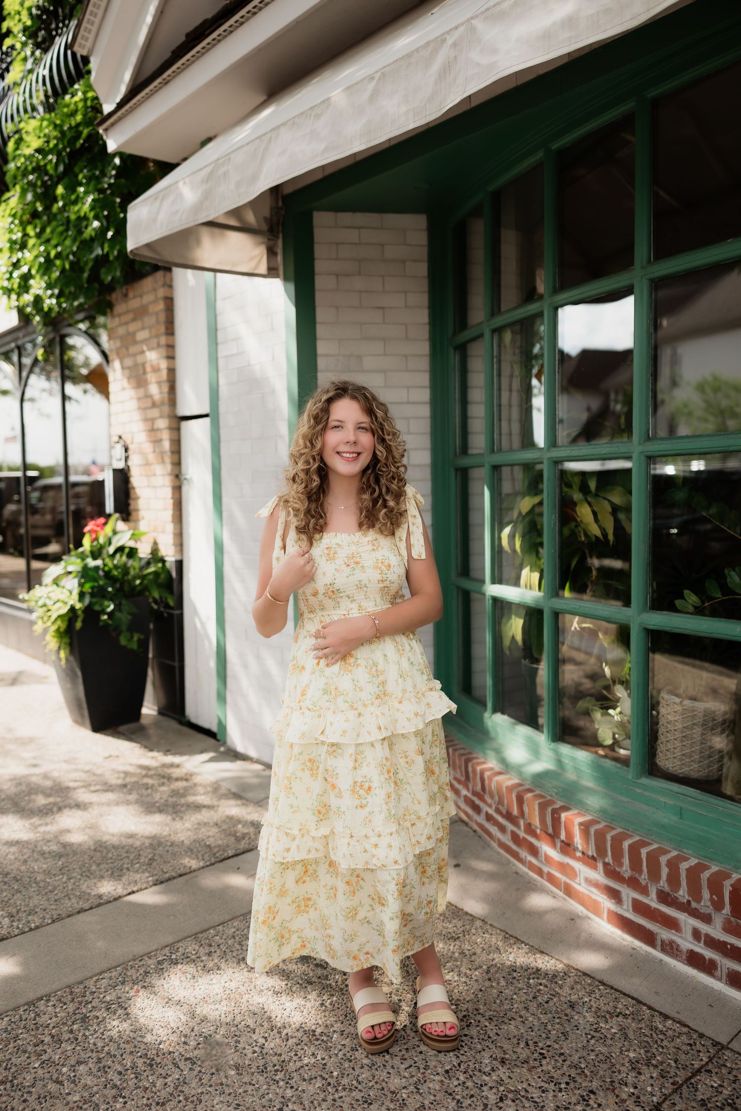 Smiling young woman wearing yellow floral maxi dress standing outside storefront with green windows.