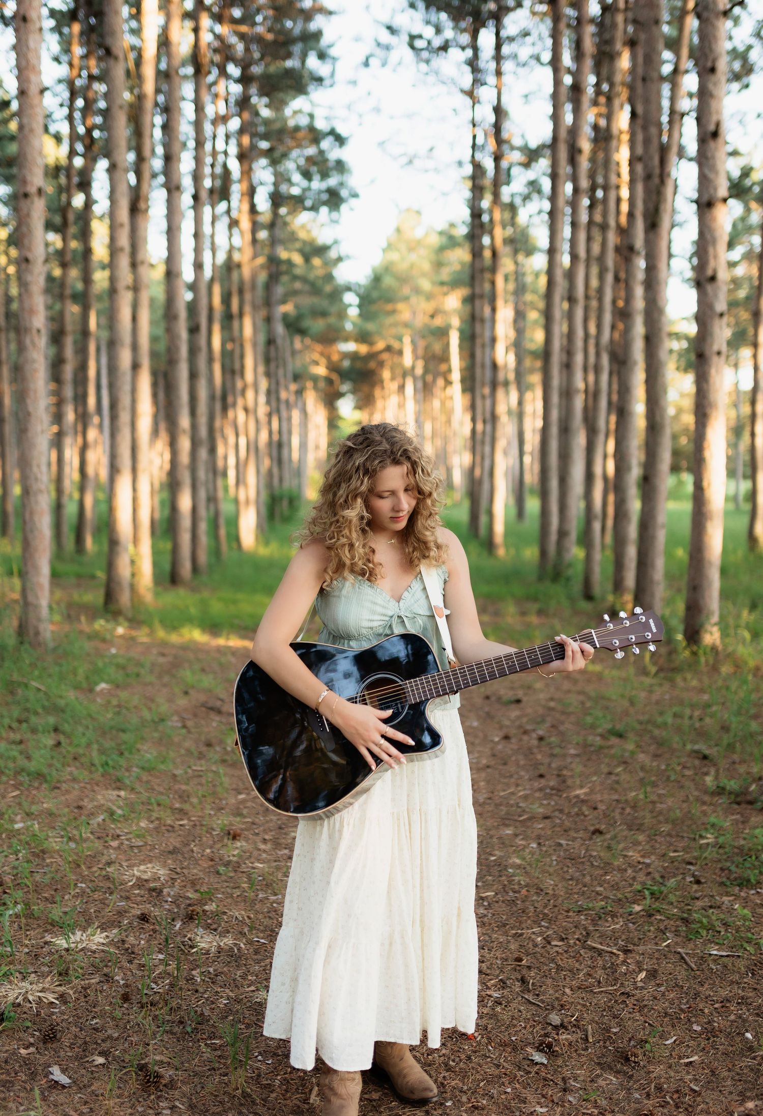 Young woman in white dress holding acoustic guitar in pine forest at sunset.