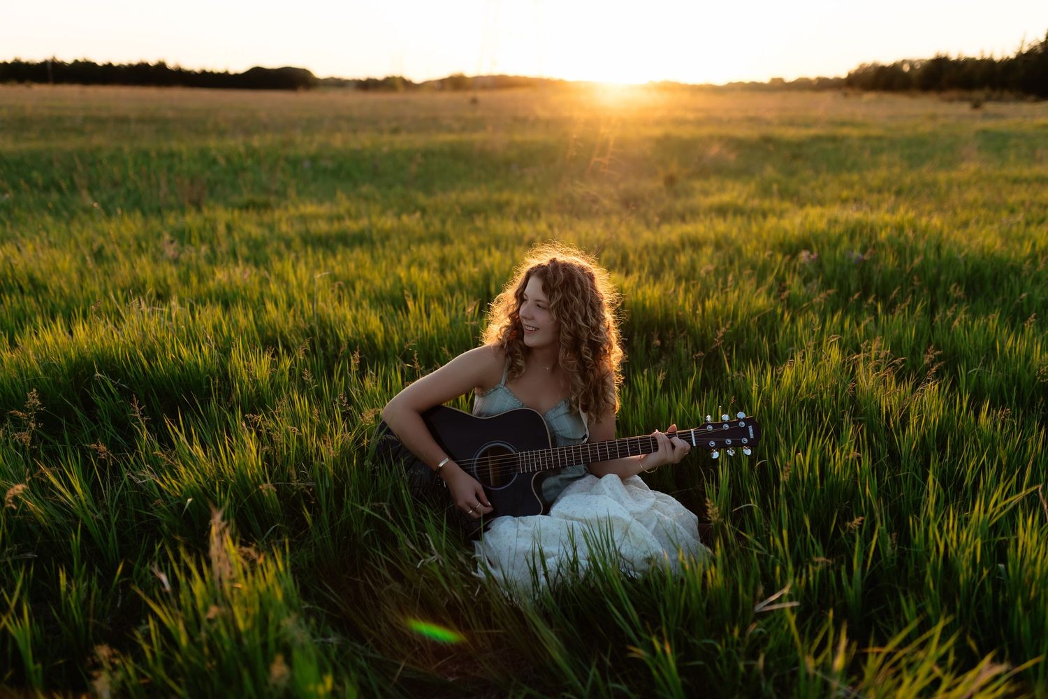 Woman playing acoustic guitar sitting in a lush green field during a golden sunset.