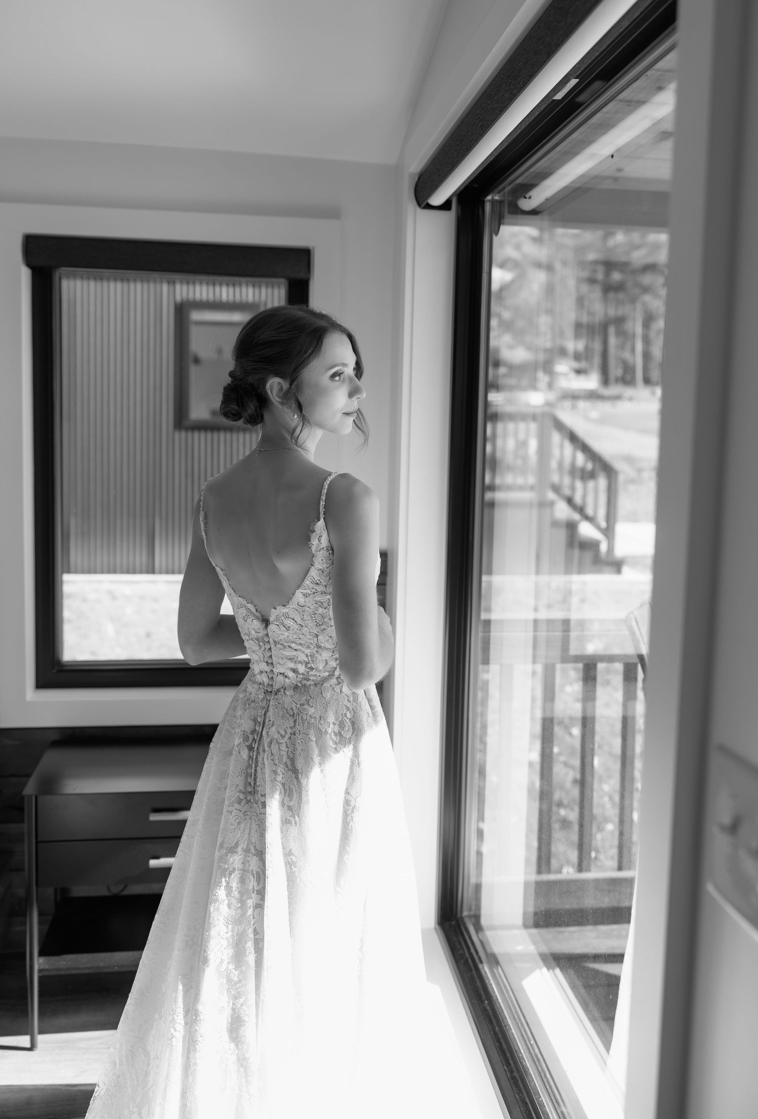 Bride in a backless lace wedding gown gazing out a sunlit window in black and white photography.