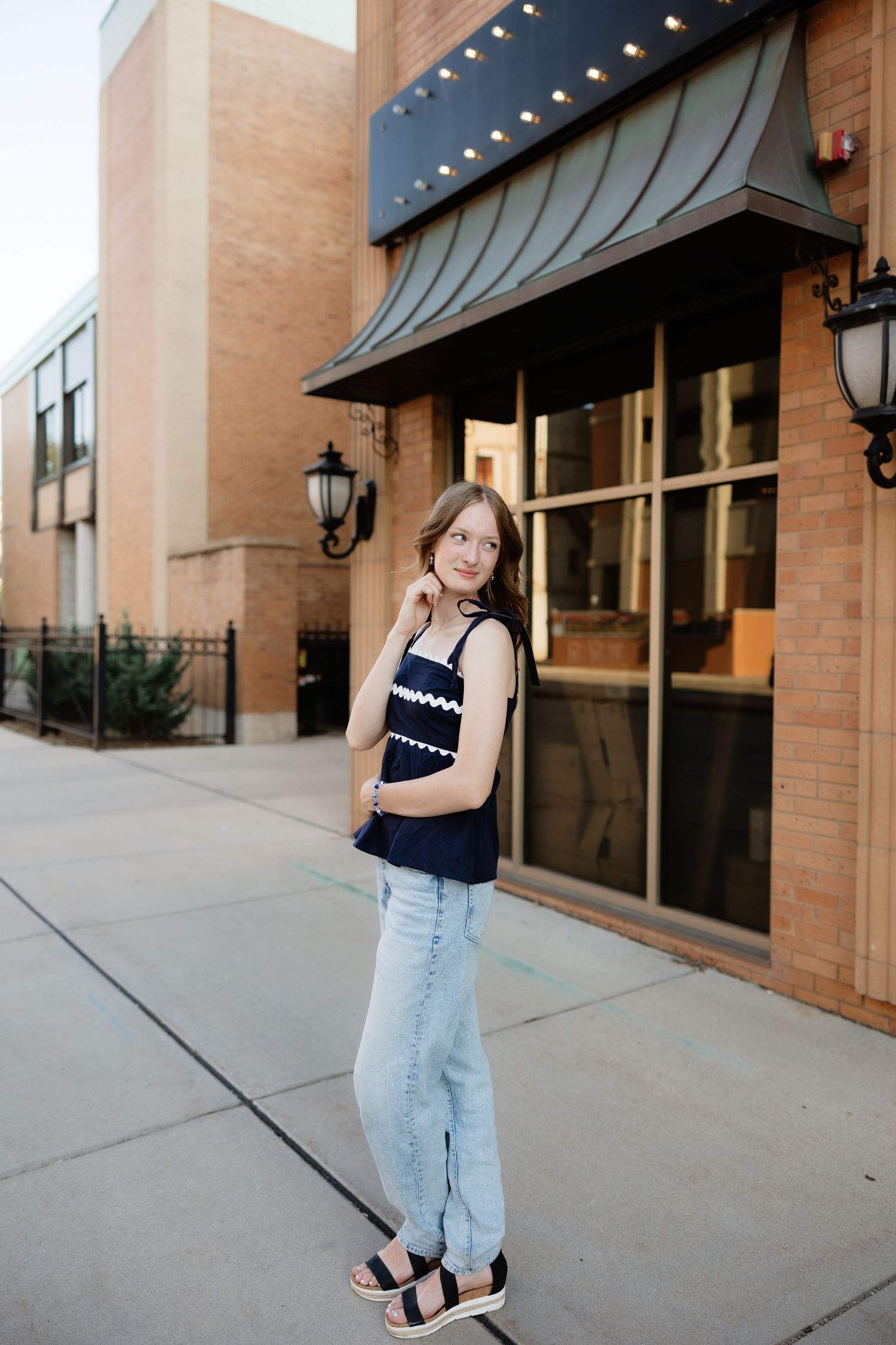 Teenage girl in navy peplum top and jeans poses outside brick building with marquee awning.