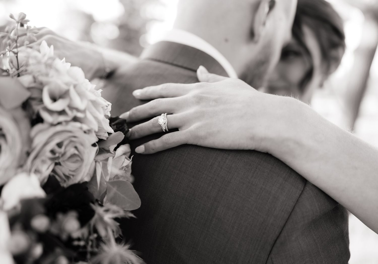 Black and white close-up of bride's diamond ring as she embraces groom holding floral bouquet.