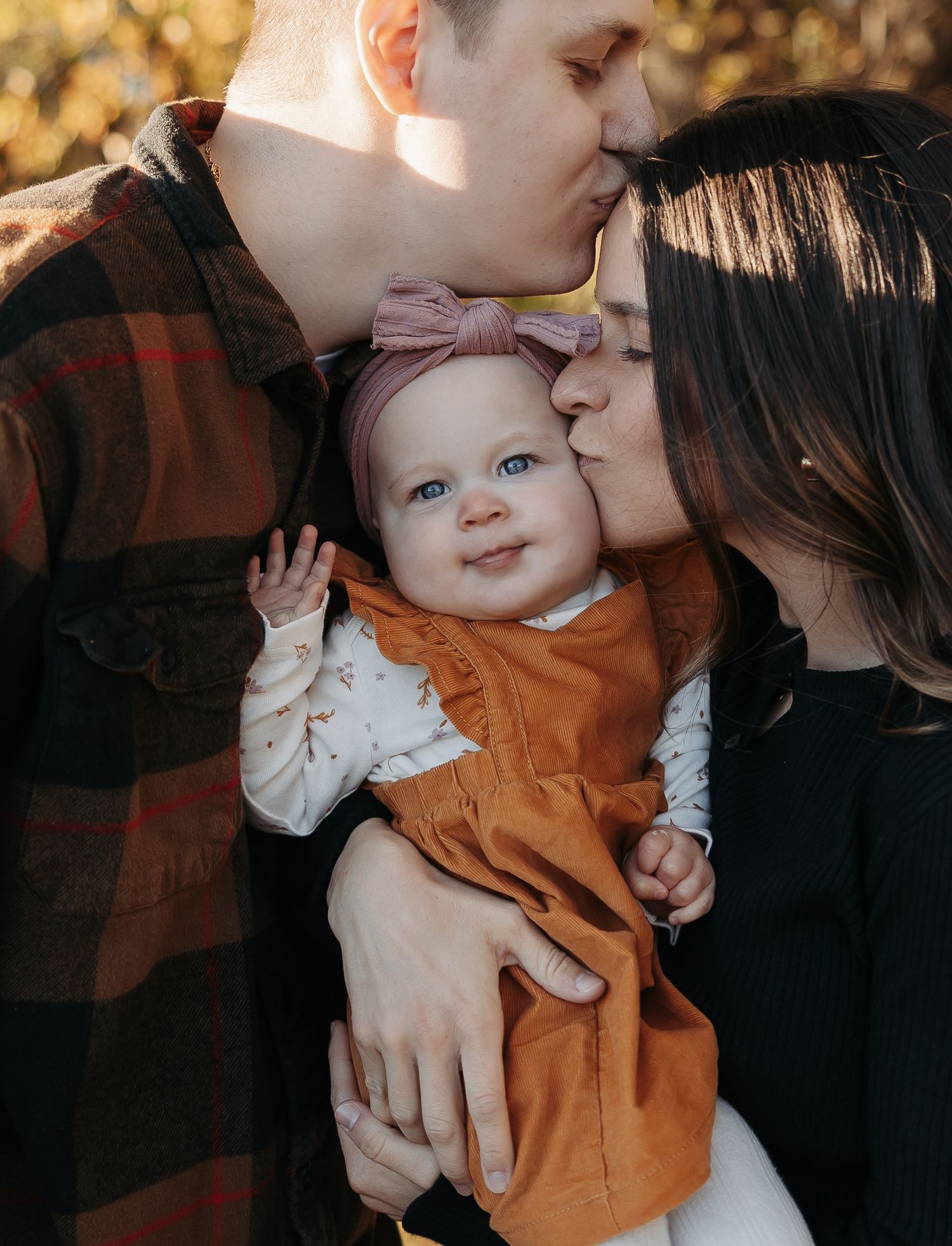 Young couple kissing their smiling baby girl in an orange dress with pink bow outdoors in autumn.