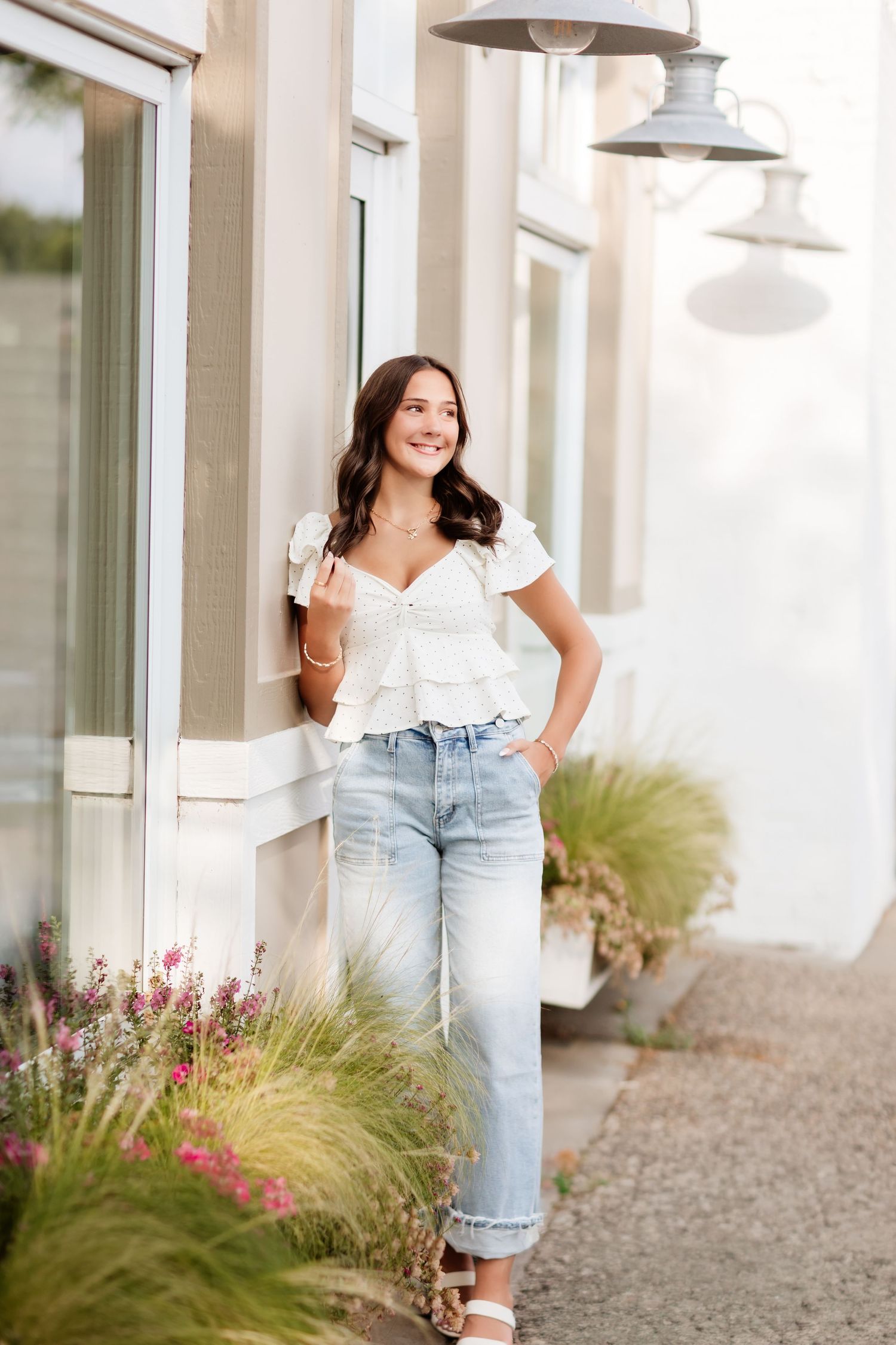 Young woman in white ruffled top and light jeans leaning against a building with hanging lights and flowers.