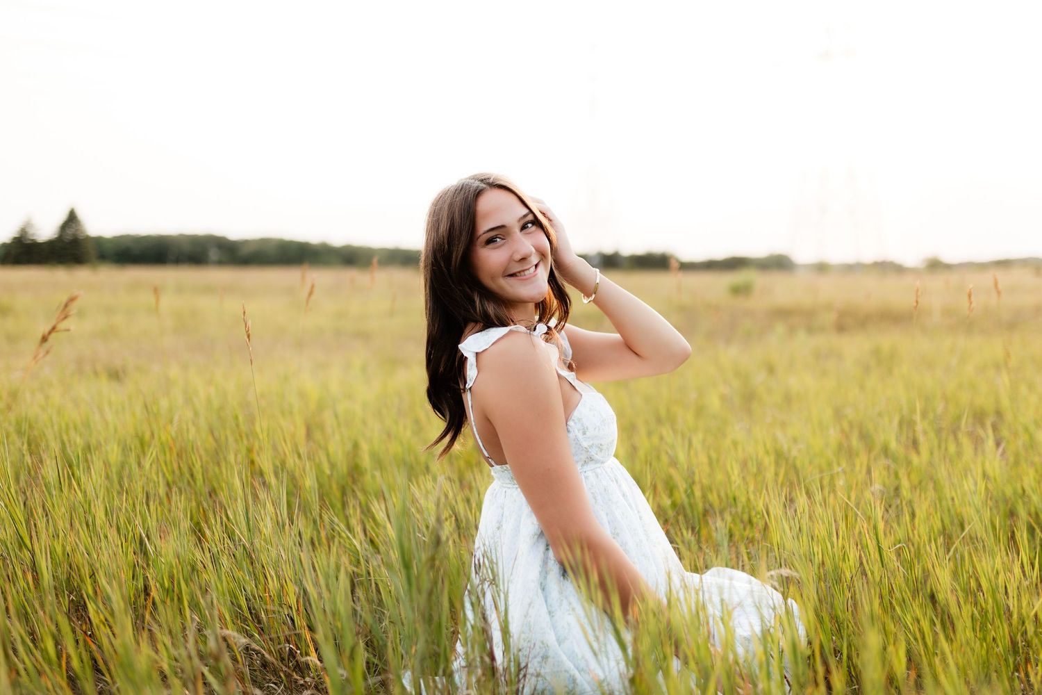 Brunette teen in white dress laughing while sitting in tall grass at sunset.