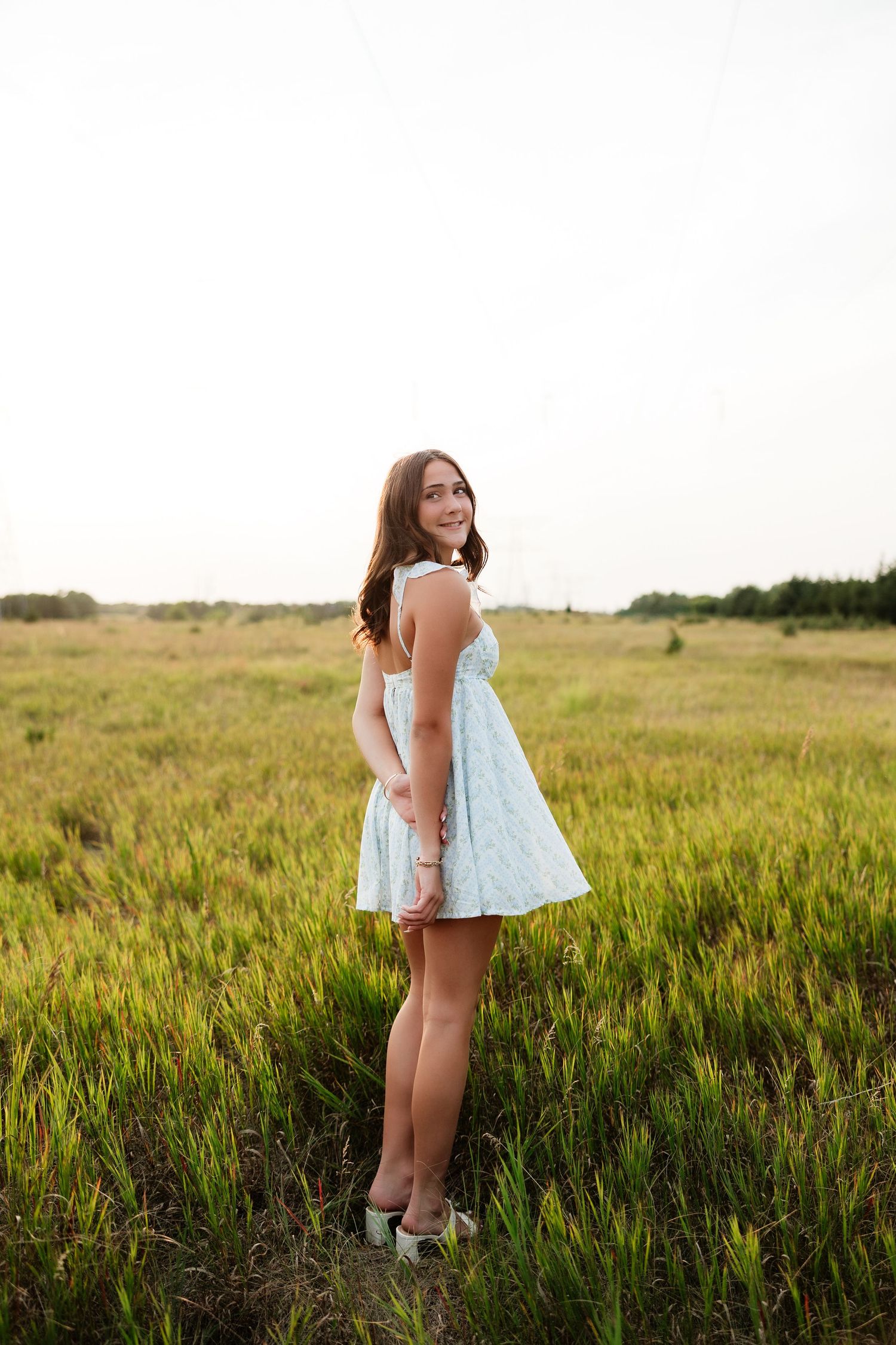 Young woman in light blue sundress standing in green field, looking back over shoulder at golden hour.