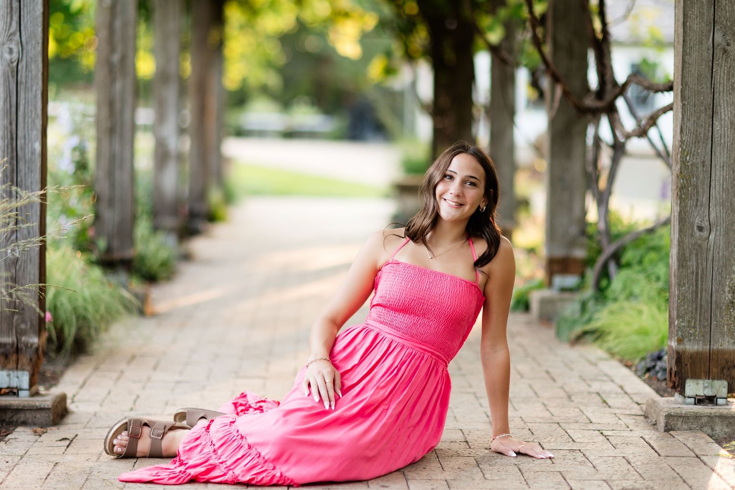 Smiling young woman in a pink maxi dress sitting on a brick pathway under a wooden pergola.