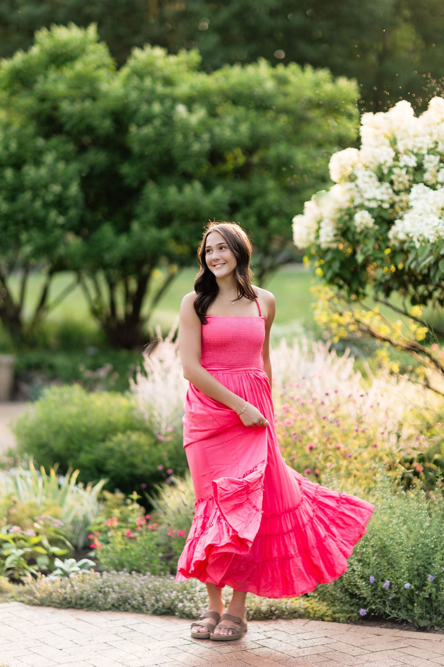 Young woman in pink maxi dress twirling in a lush garden with white hydrangeas.