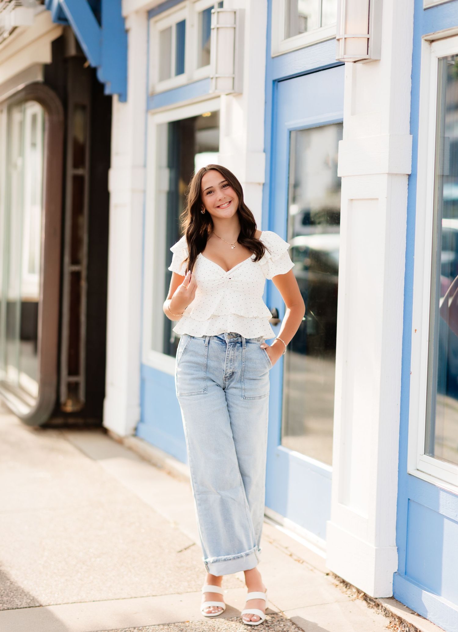 Smiling brunette woman in white blouse and light jeans posing on sidewalk by blue building.