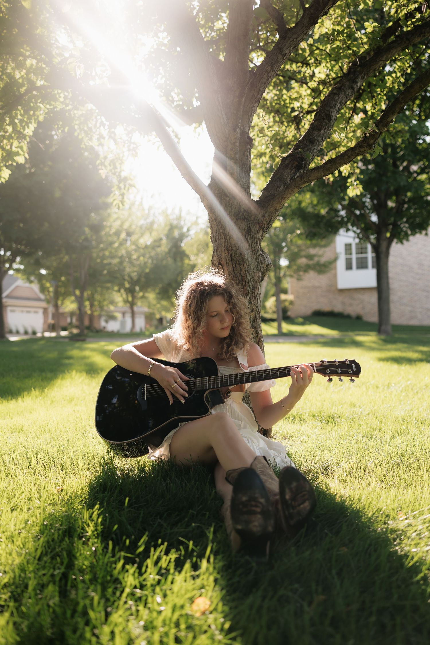 Young woman with curly hair playing black acoustic guitar while sitting on green grass under a tree in sunlight.