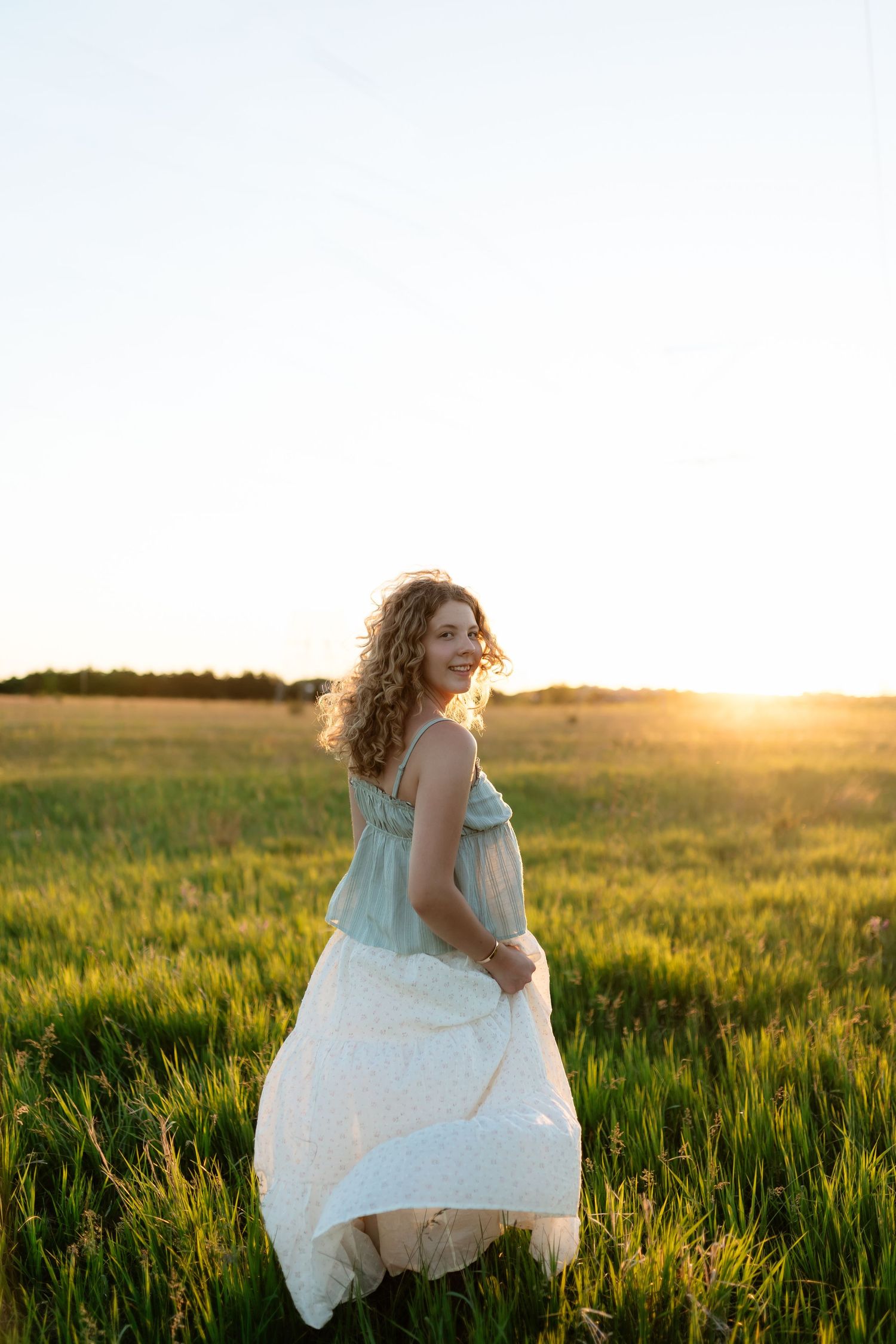 Young woman in mint and white dress walking through a golden sunlit field at sunset.