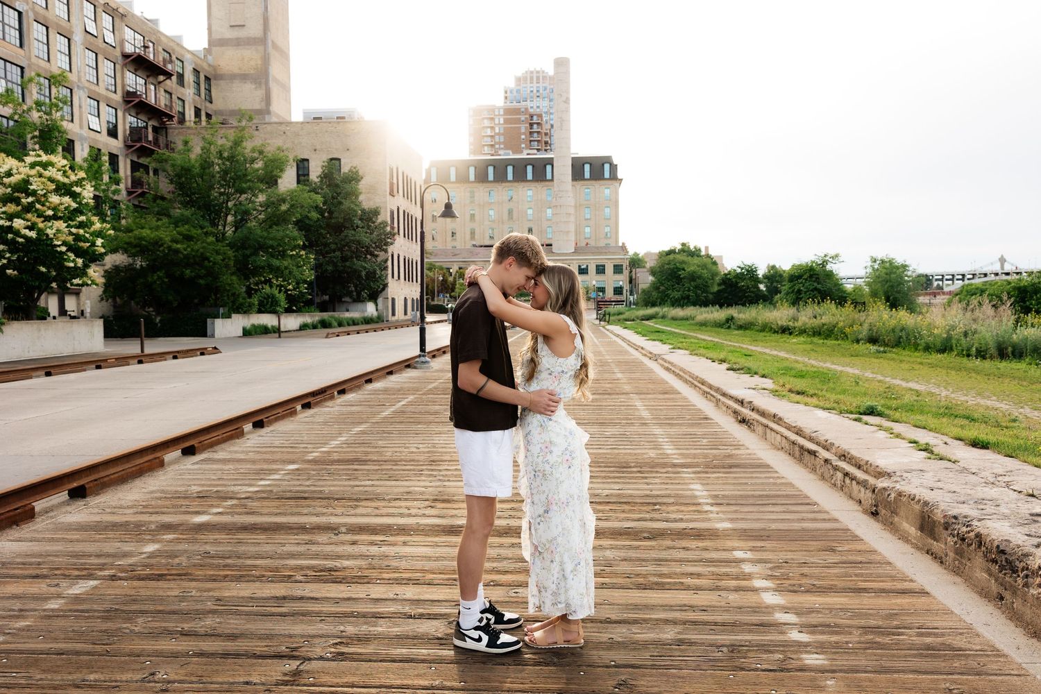 Couple sharing a romantic embrace on a wooden boardwalk with urban buildings in the background on a sunny day.