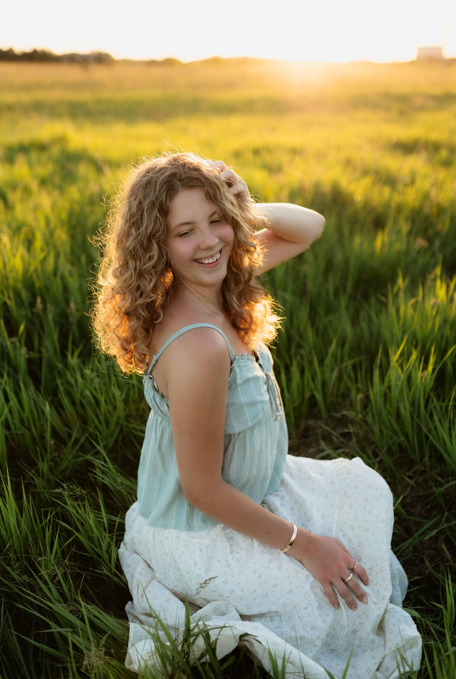 Young woman with curly hair in a mint sundress sits smiling in a green field bathed in golden sunset light.