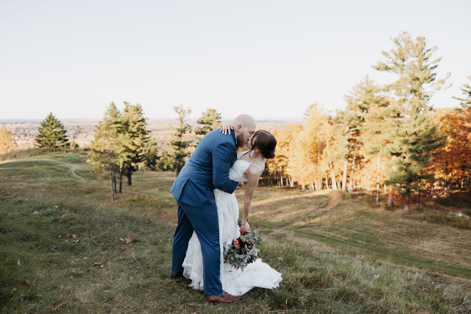 Groom dipping bride for a kiss on a golden autumn hillside with colorful fall foliage behind them.