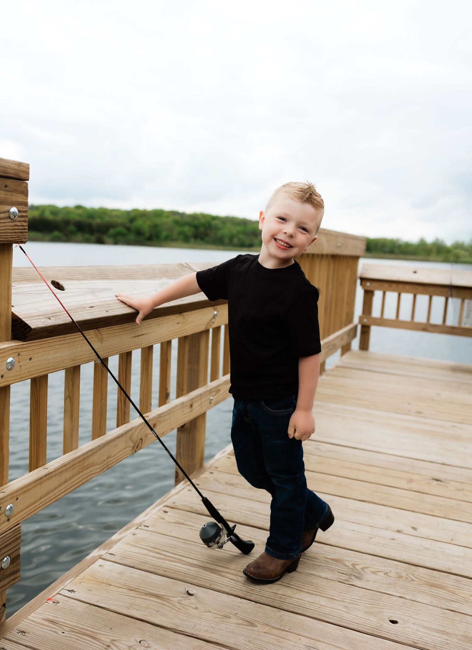 Smiling young boy holding a fishing rod on a wooden dock by a lake.
