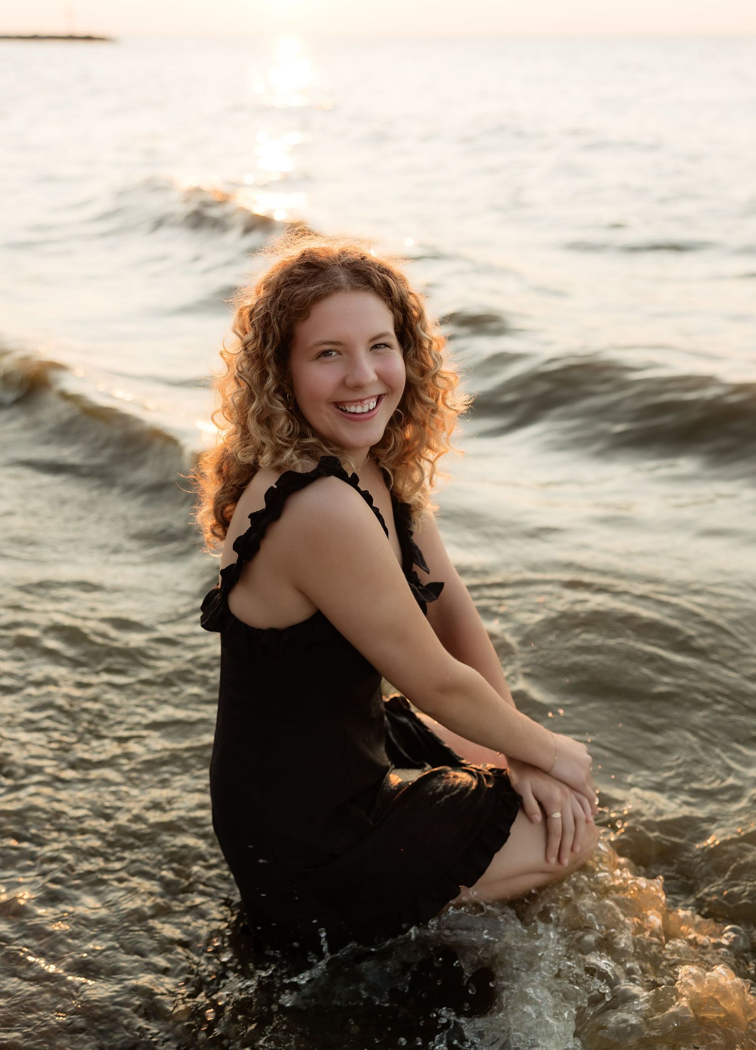 A smiling woman with curly red hair sits in ocean waves wearing a black dress at golden hour sunset.
