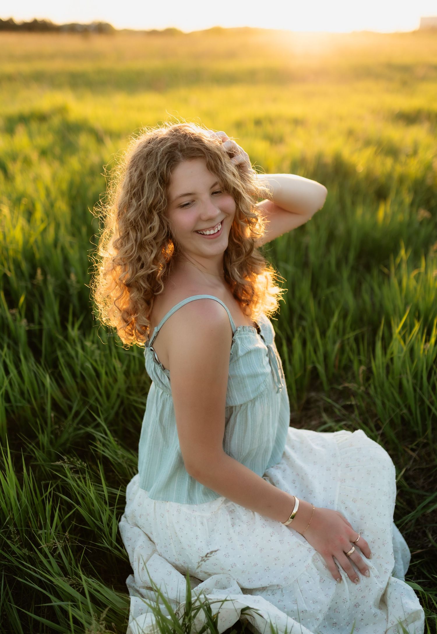 Smiling young woman with curly hair in a blue sundress sitting in a sunlit green field at sunset.