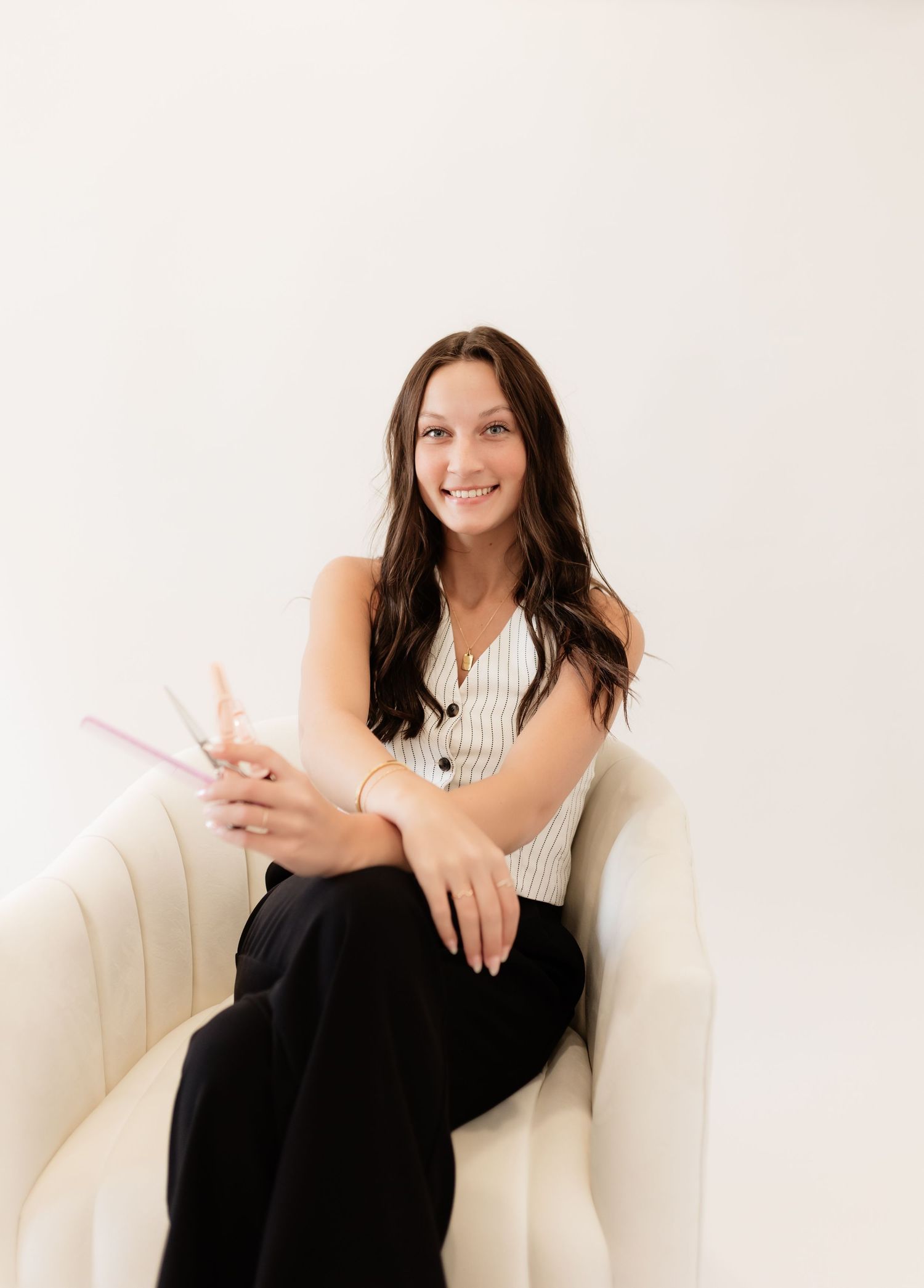 Young brunette woman sitting in a cream chair smiling while holding pink hair scissors in a bright studio.