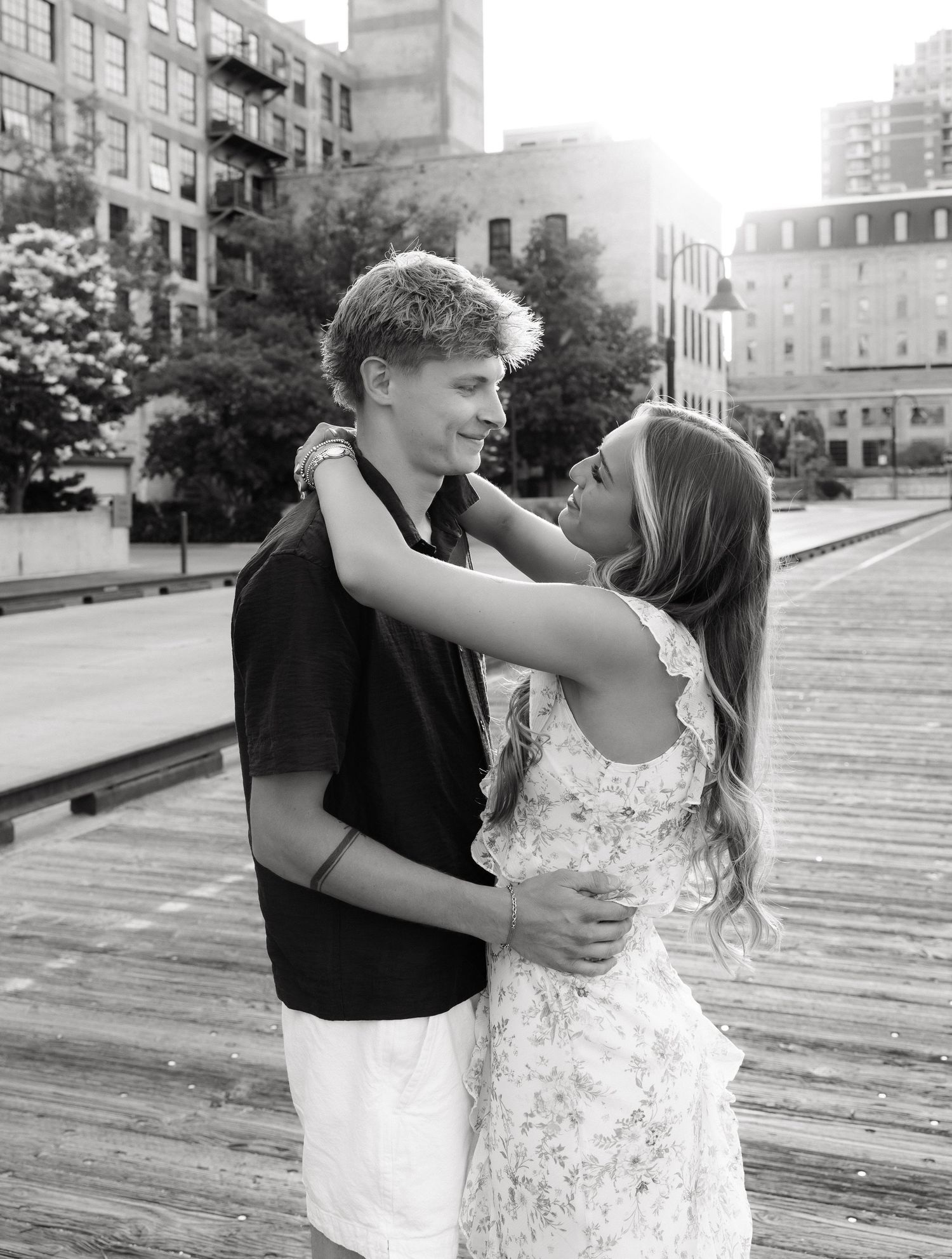 Black and white photo of a couple embracing and smiling at each other on a wooden boardwalk with city buildings behind them.