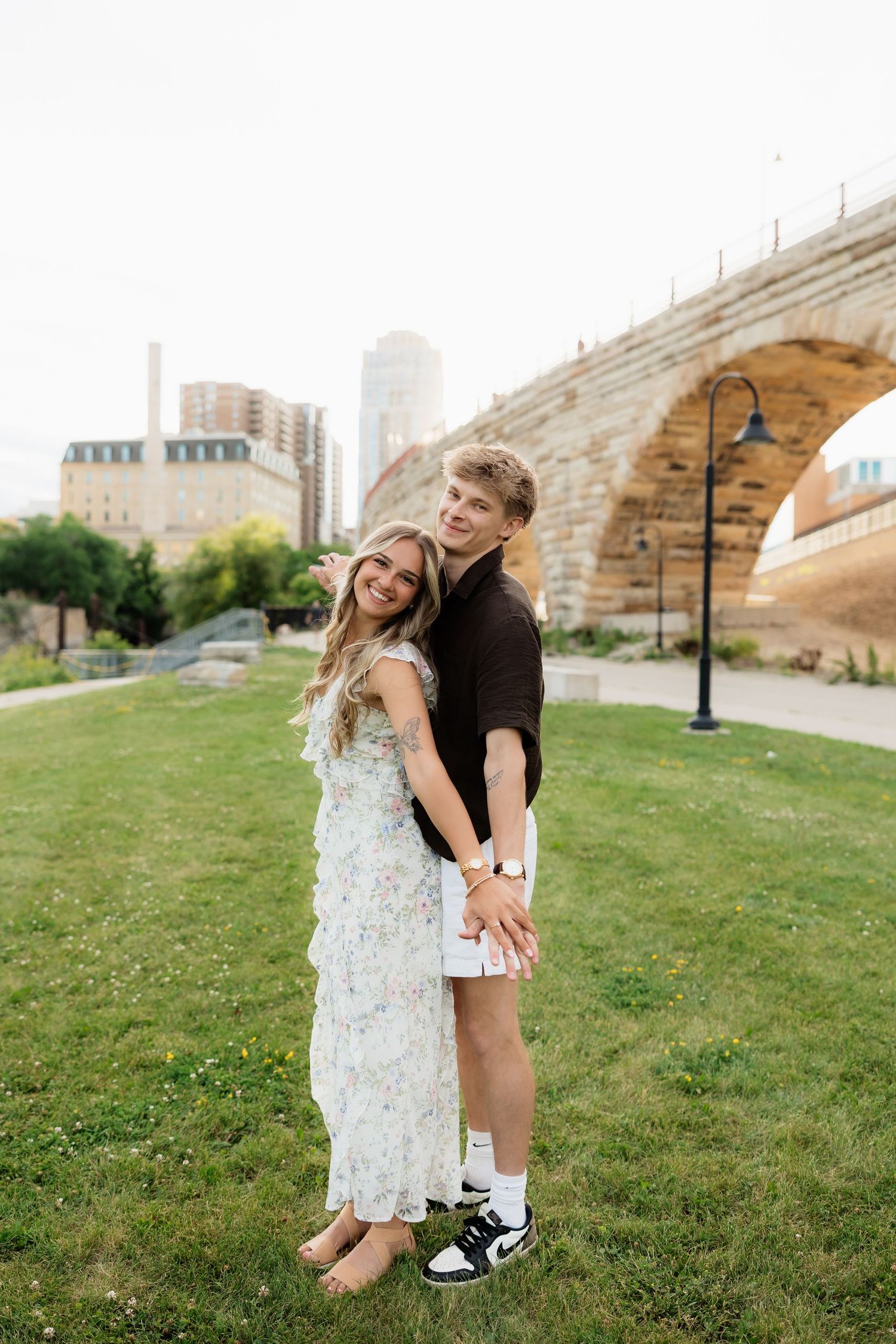 Couple holding hands and smiling on green grass with a classic stone arch bridge and city skyline behind them.