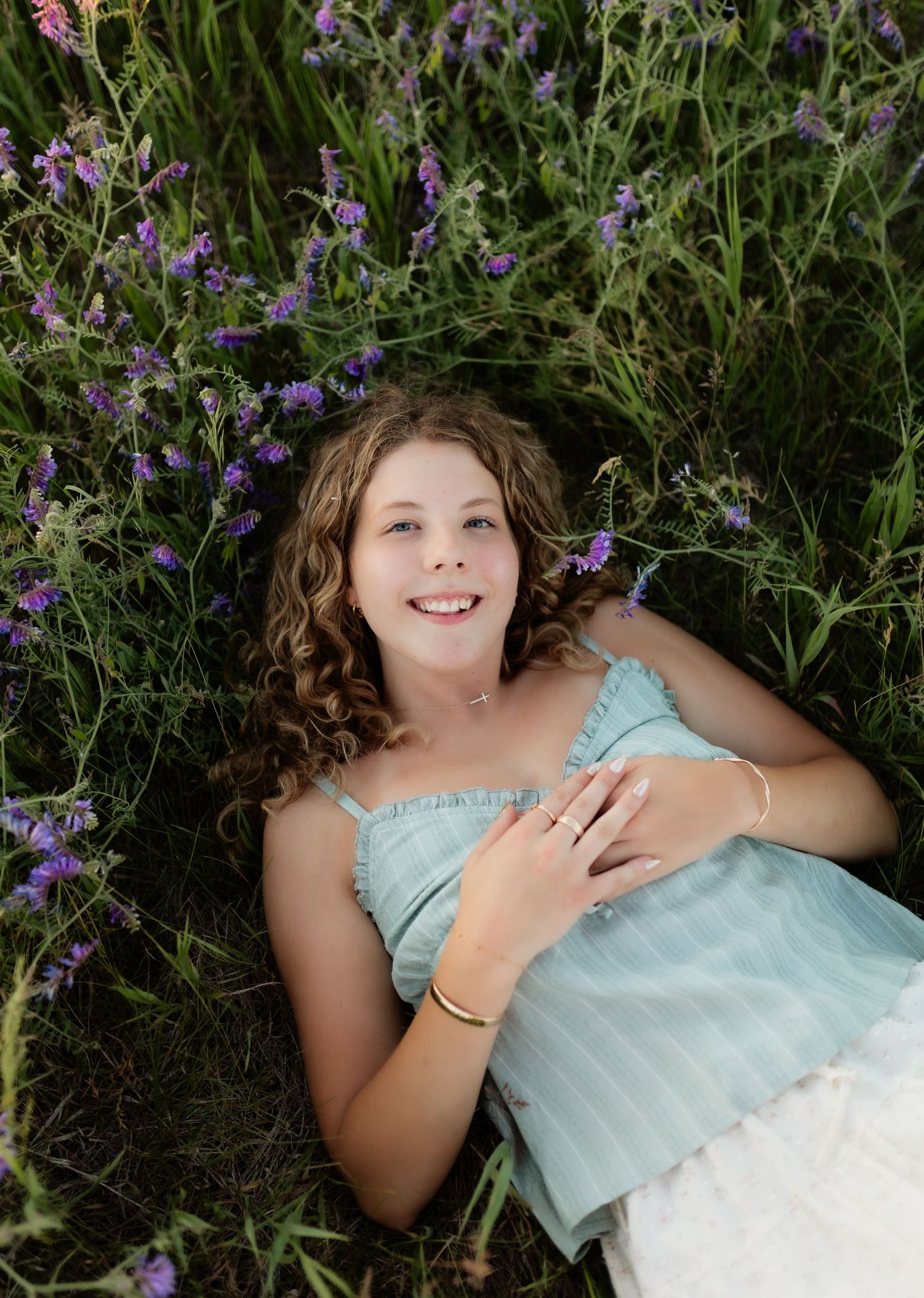 Young woman with curly hair lying among purple wildflowers in a green field wearing a light blue dress.
