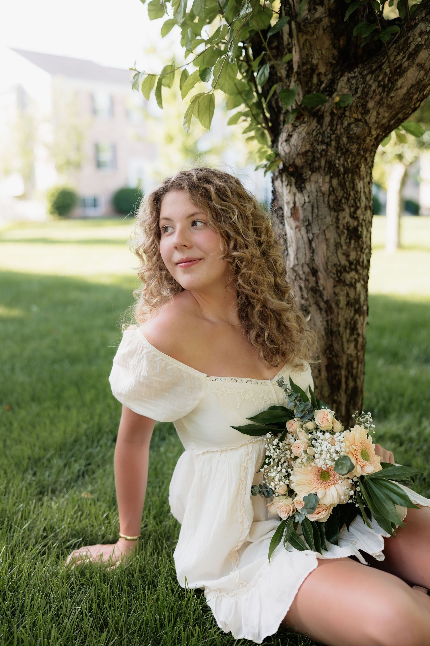 Young woman with curly hair in white dress sitting on grass under a tree holding a floral bouquet.