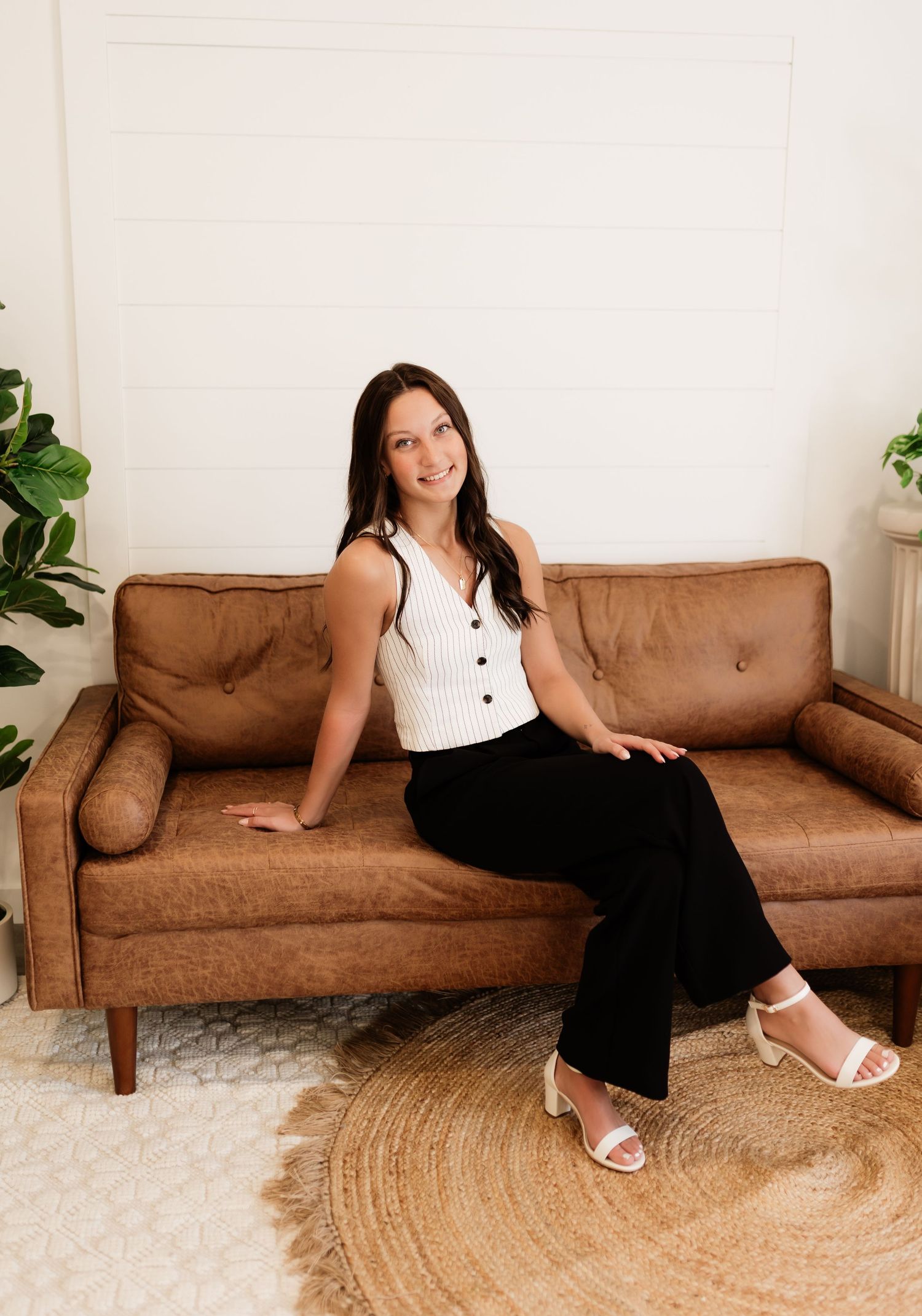 Woman in white top and black pants sitting on brown leather sofa smiling.
