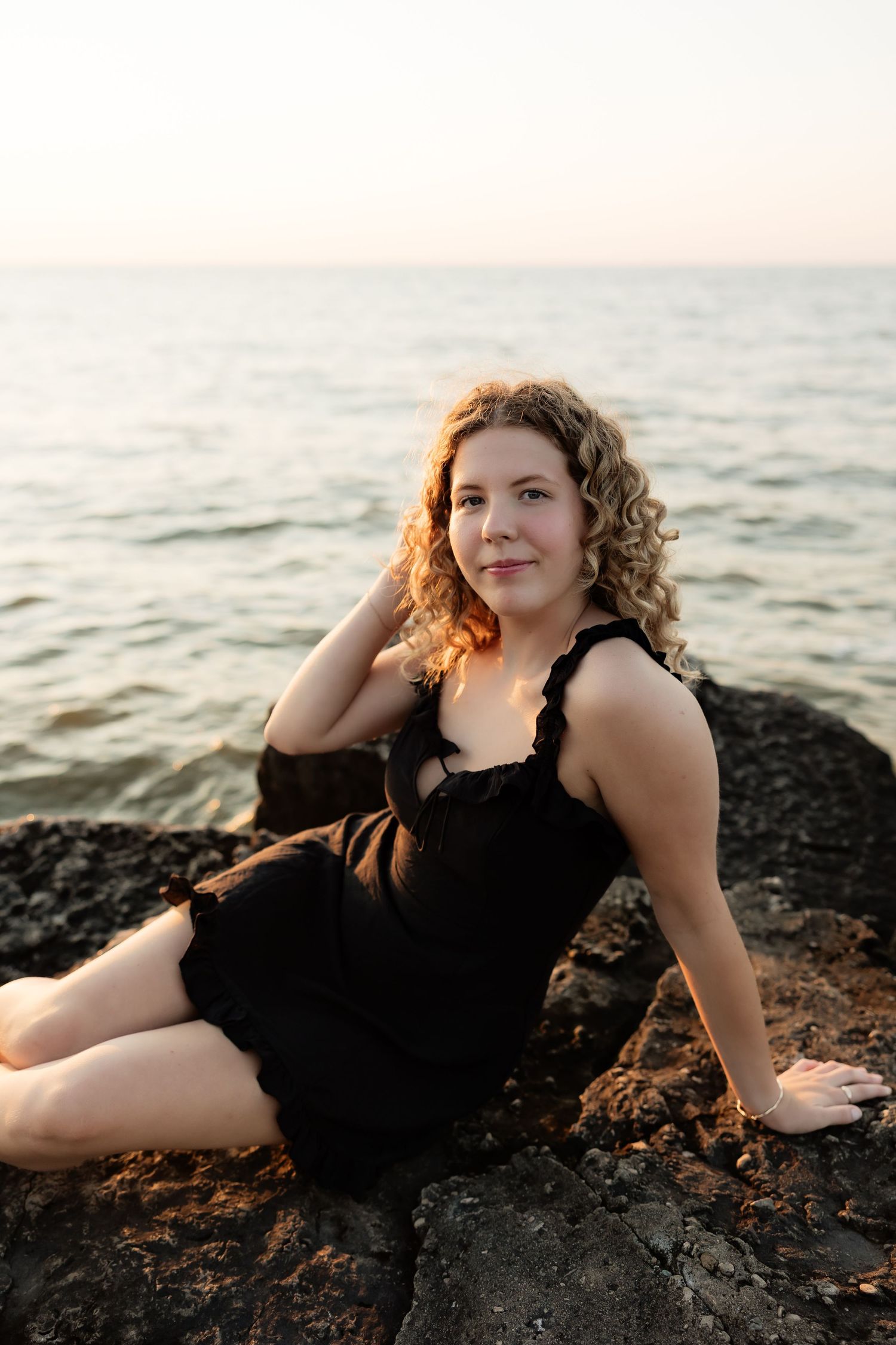 Woman in black dress sitting on rocky shore at sunset with calm ocean in background.