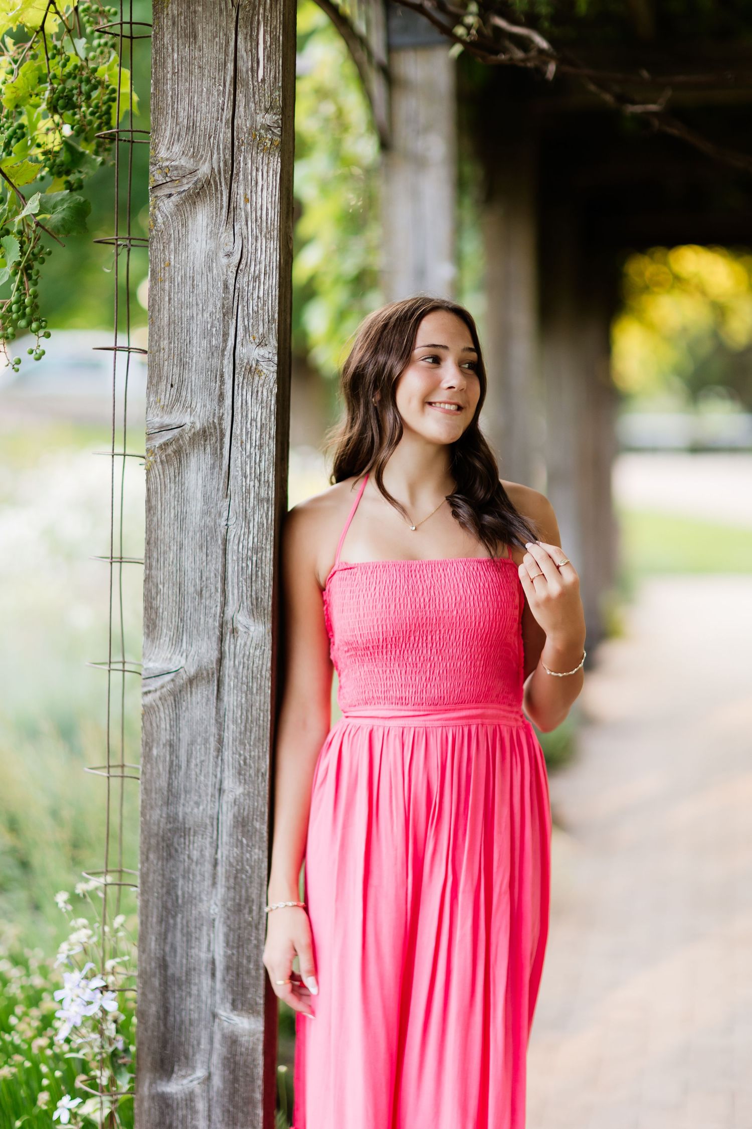 Young woman in bright pink maxi dress gazing sideways while leaning on a wooden post in a garden.
