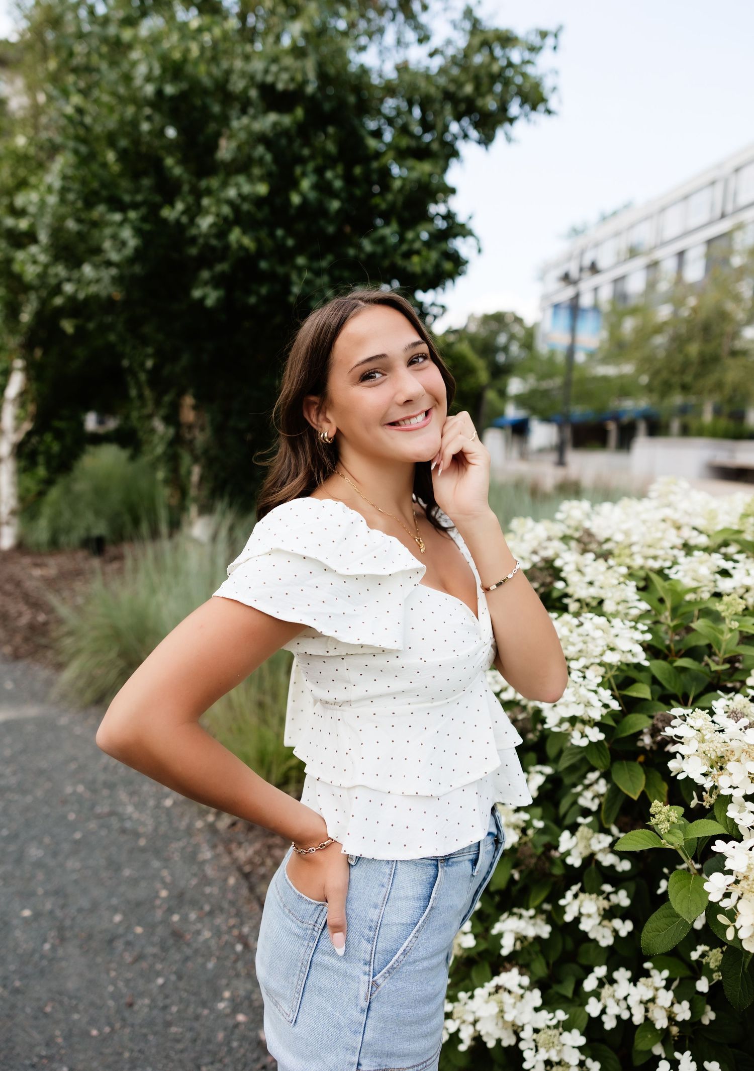 Young woman in white ruffled top and jeans smiling beside white flowering bushes in an urban garden.
