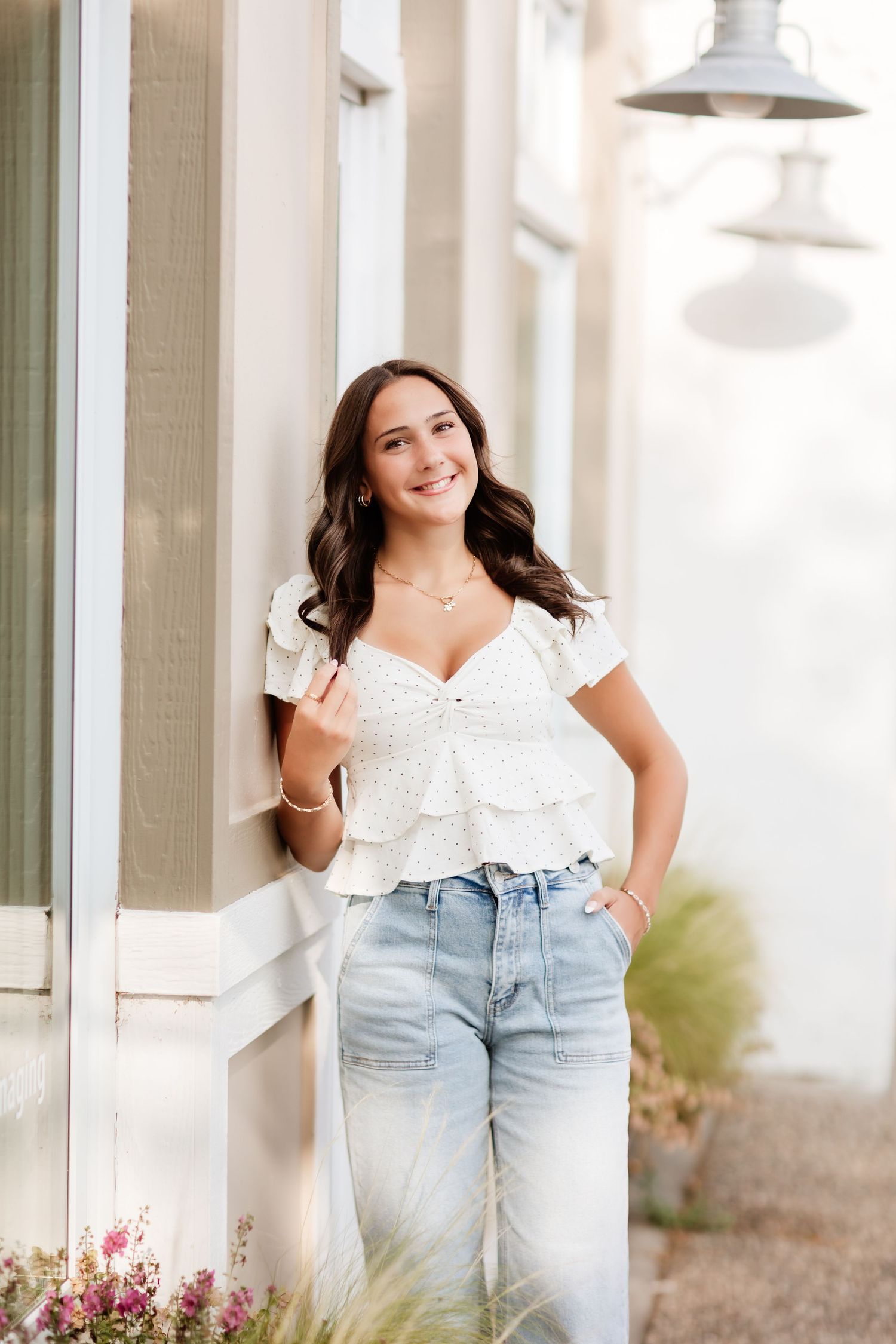 Young woman in white ruffled top and jeans leaning against a building exterior, smiling outdoors.
