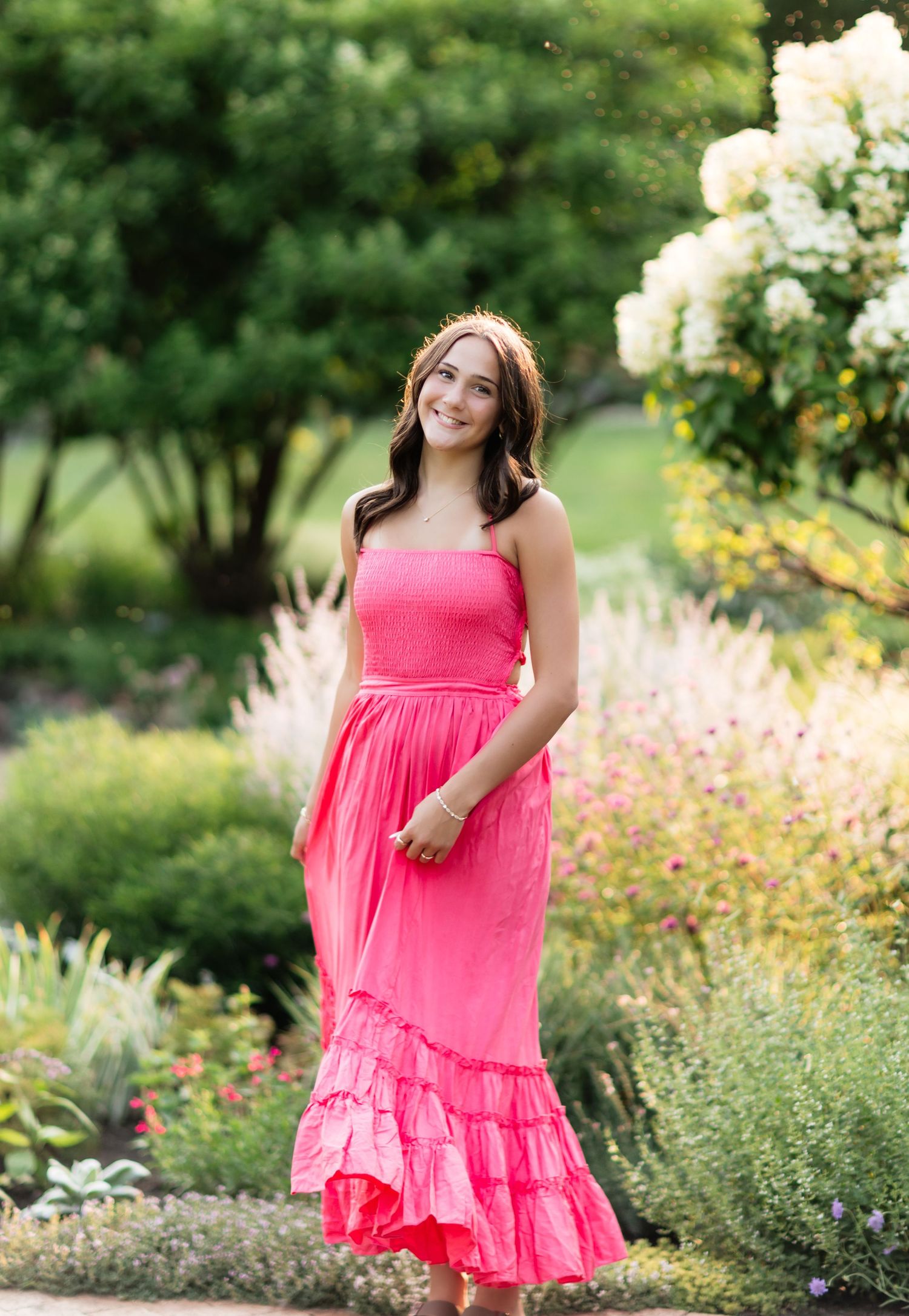 Young woman in bright coral strapless ruffle maxi dress walking through lush garden in summer.