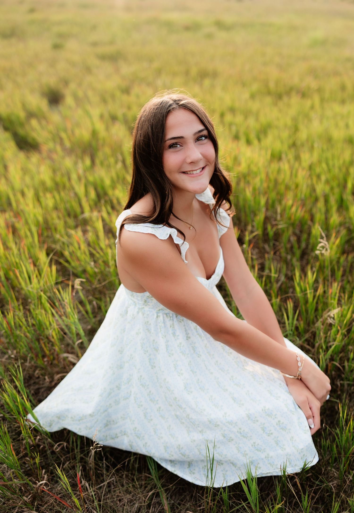 Smiling teen girl in white sundress sitting in tall green grass field at golden hour.