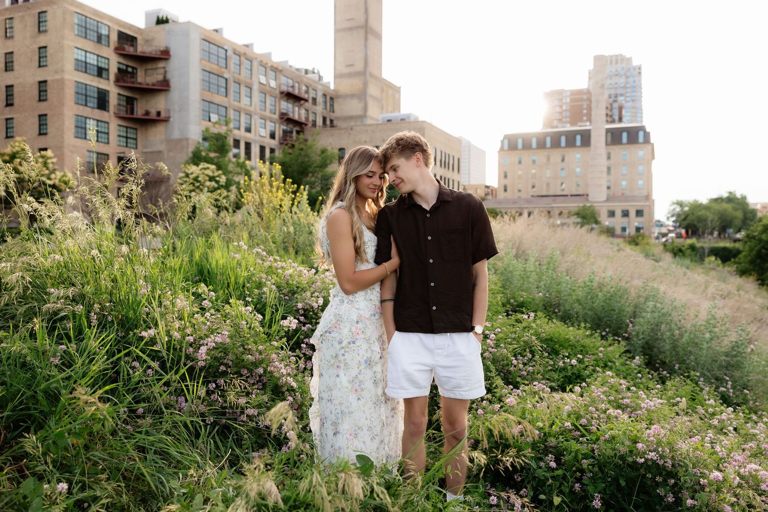 Young couple standing together smiling among wildflowers with urban buildings visible in the background.
