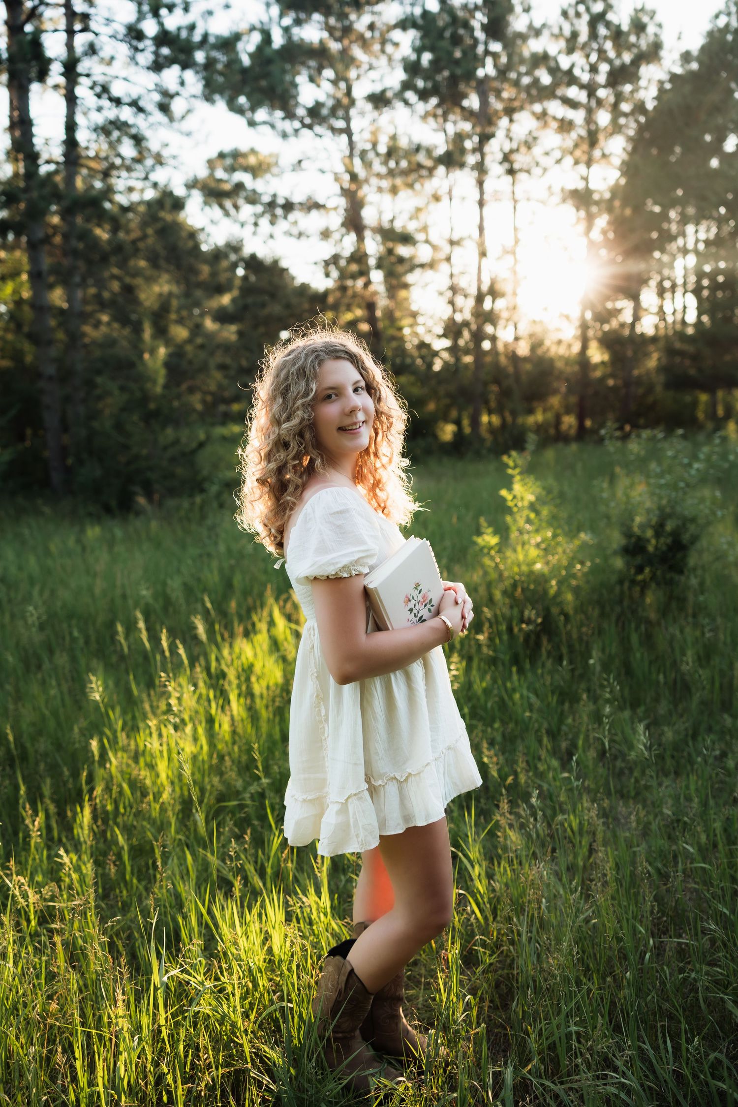 Young woman in white dress and cowboy boots holding a book in a sunlit green meadow with forest background.