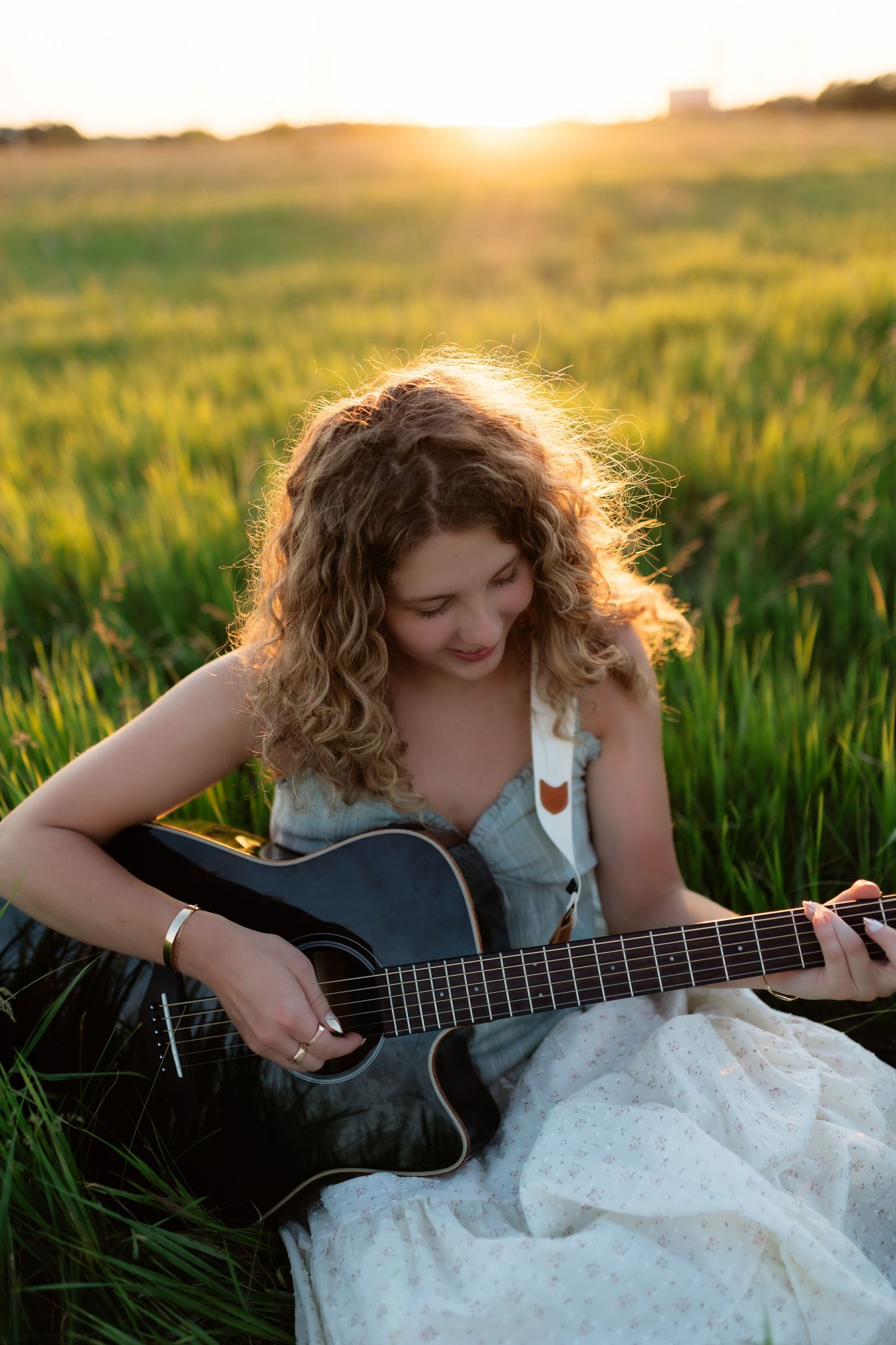 Young woman with curly hair smiling while playing black acoustic guitar sitting in a green field at golden sunset.
