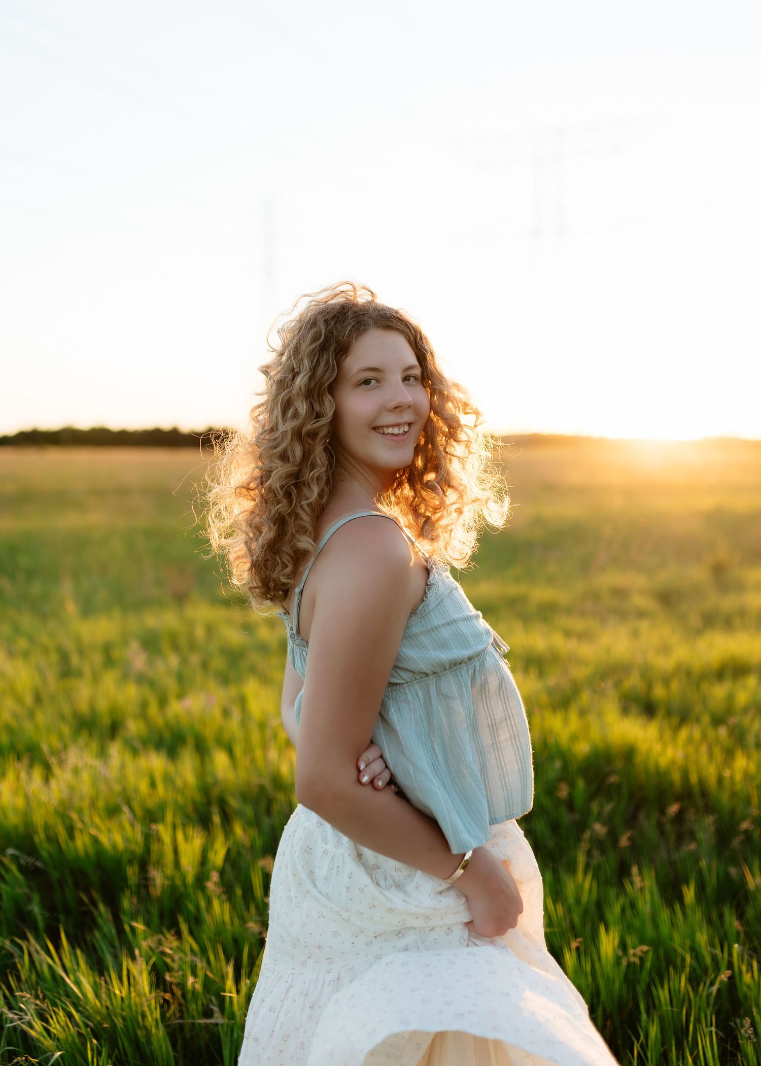 Smiling pregnant woman with curly hair in a light blue top and white skirt standing in a sunlit green field.
