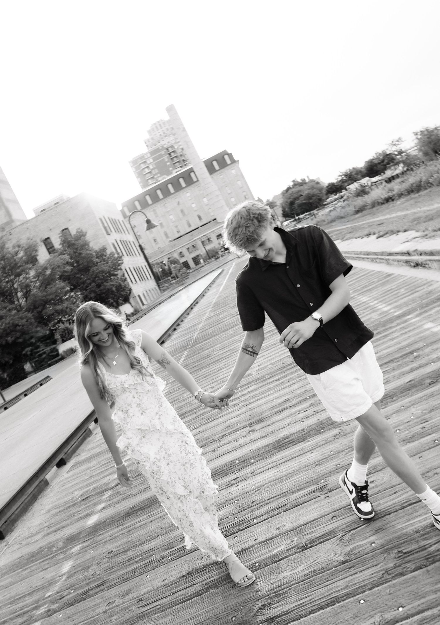 Black and white photo of a young couple holding hands and dancing on a wooden boardwalk with city buildings in the background.