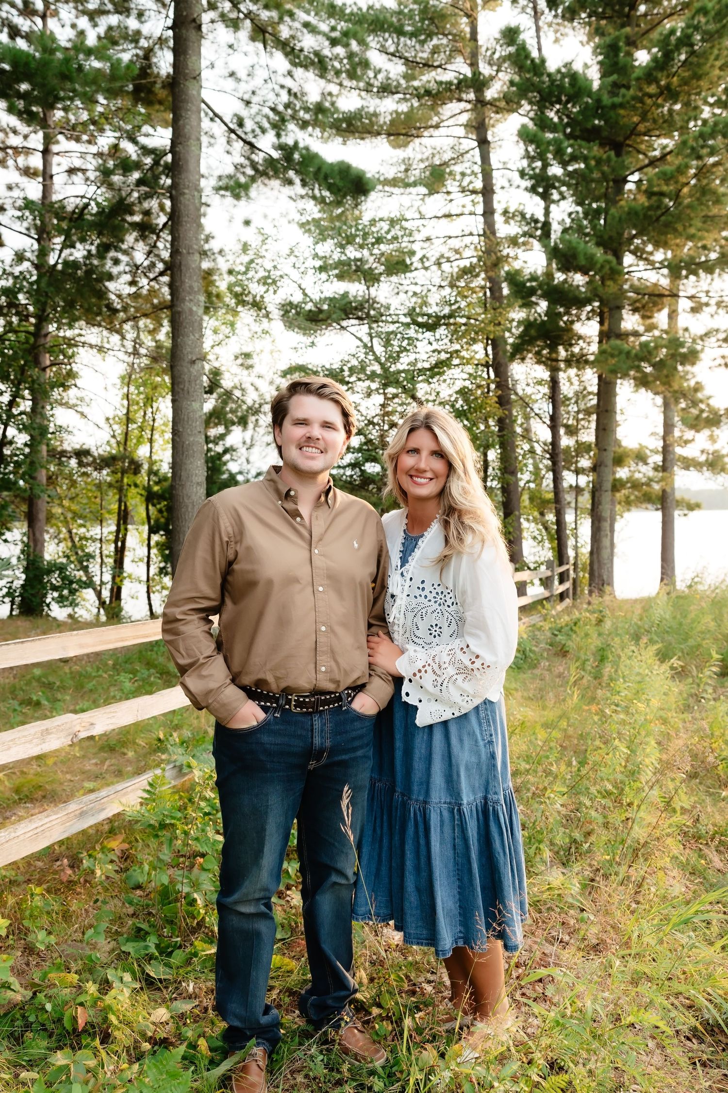 A smiling couple poses outdoors among tall pine trees near a wooden fence, dressed in casual country-style attire.