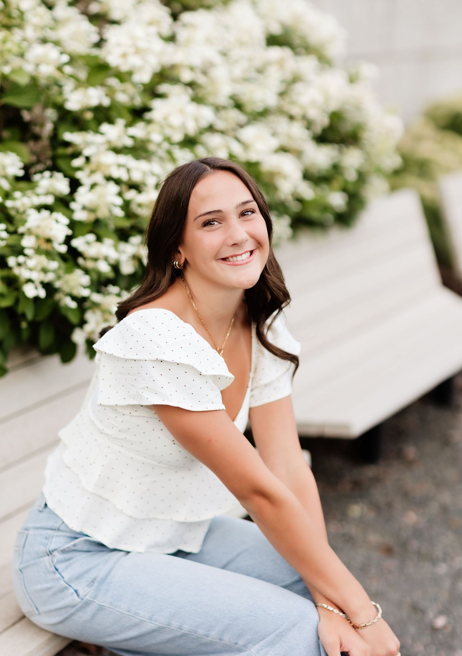 Smiling brunette woman in white off-shoulder top and jeans posing near white flowering bushes.