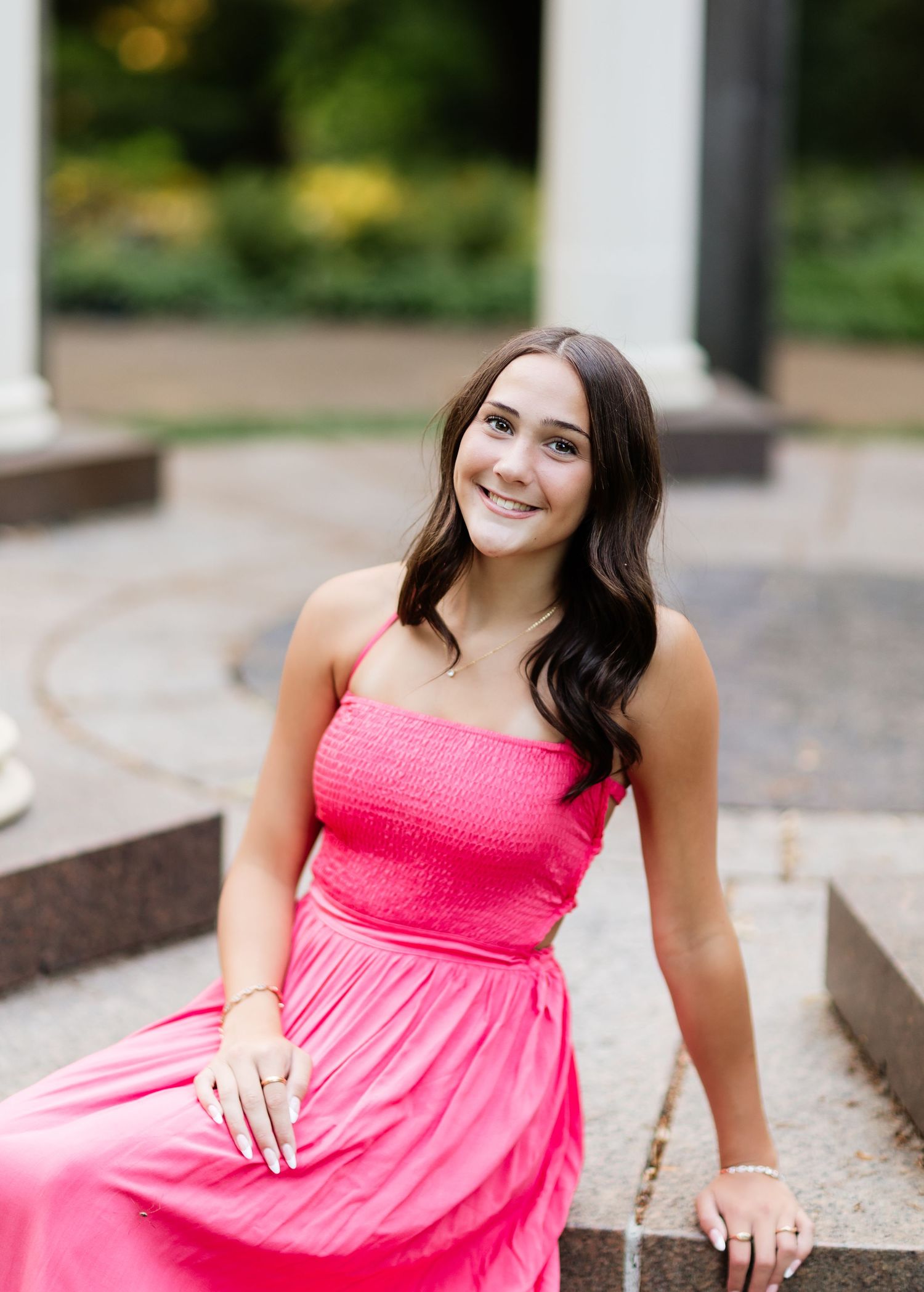 Smiling brunette in hot pink dress seated on stone steps outdoors.