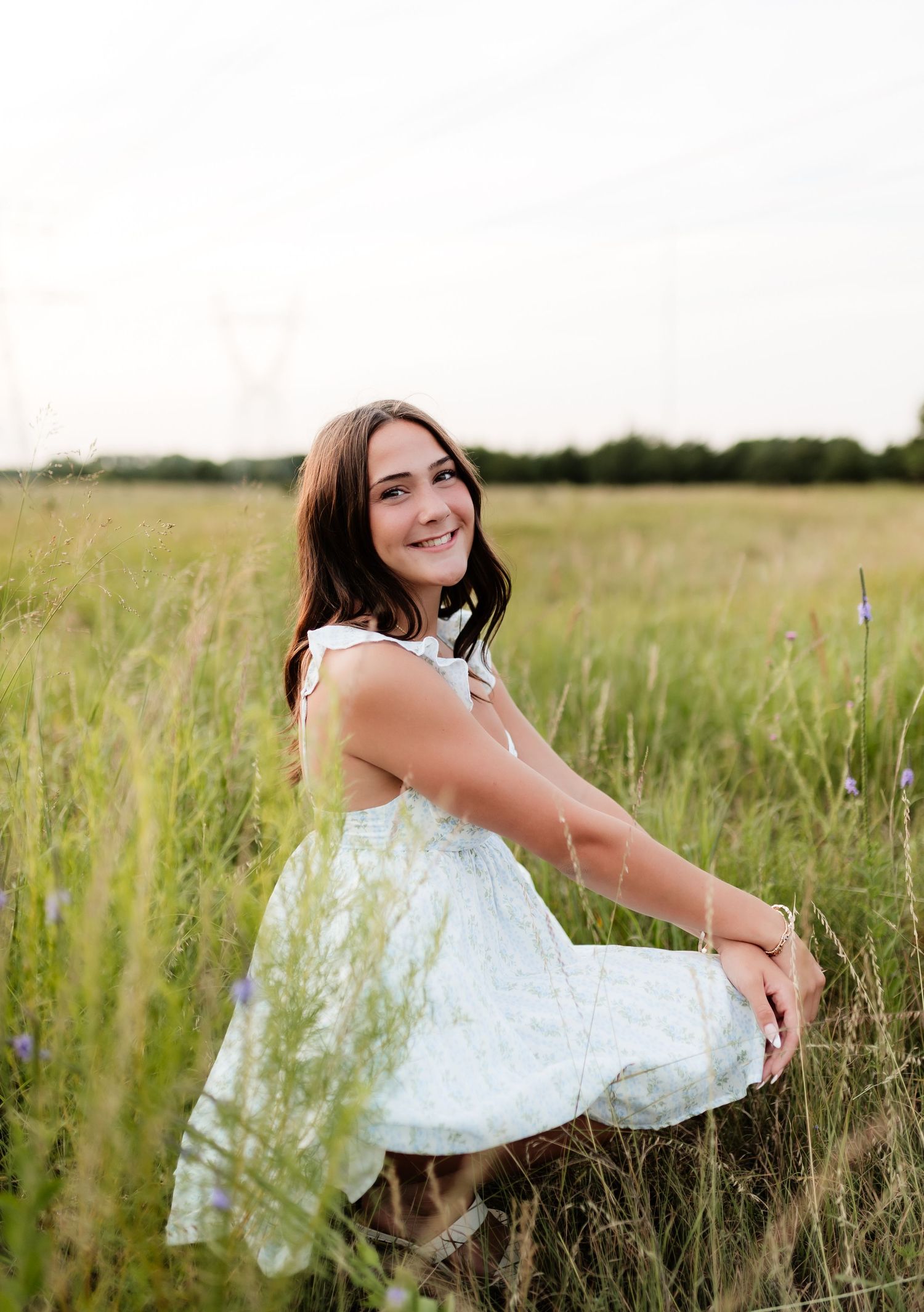 Young woman in white sundress smiling while seated in tall grass field with wildflowers.