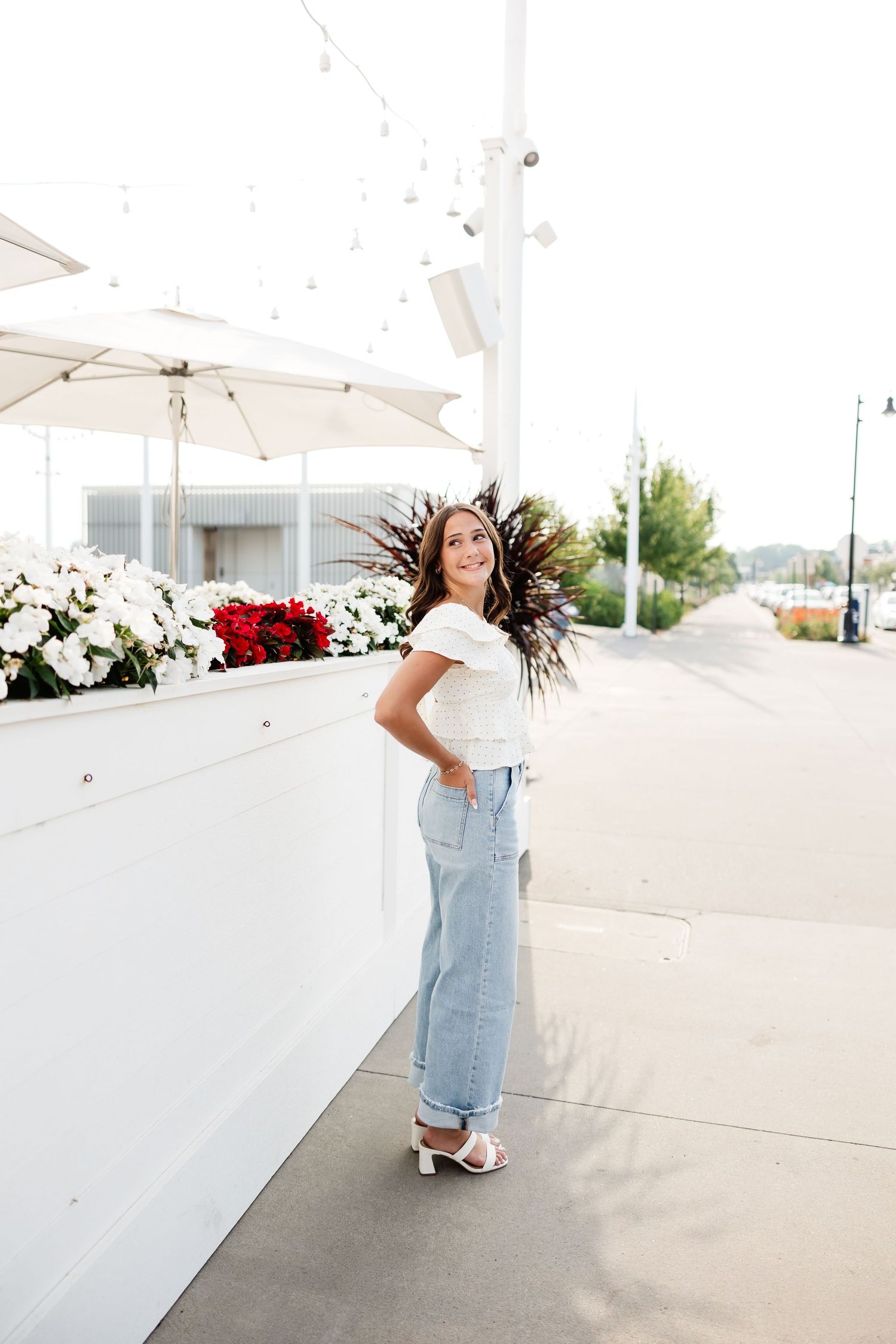 Young woman in white top and jeans posing outdoors near white planters with colorful flowers.