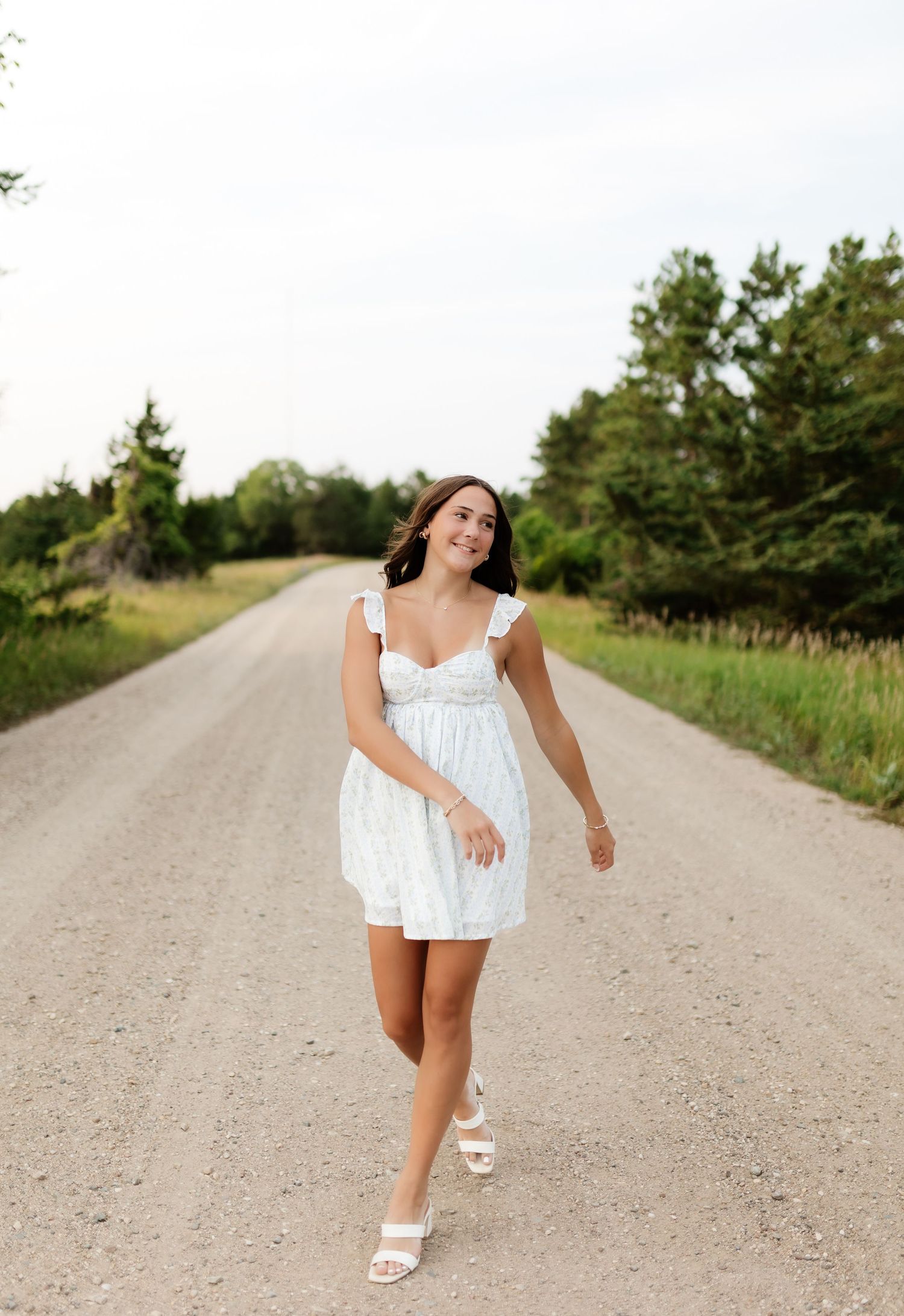 Young woman in white mini dress walking on rural dirt road surrounded by trees.