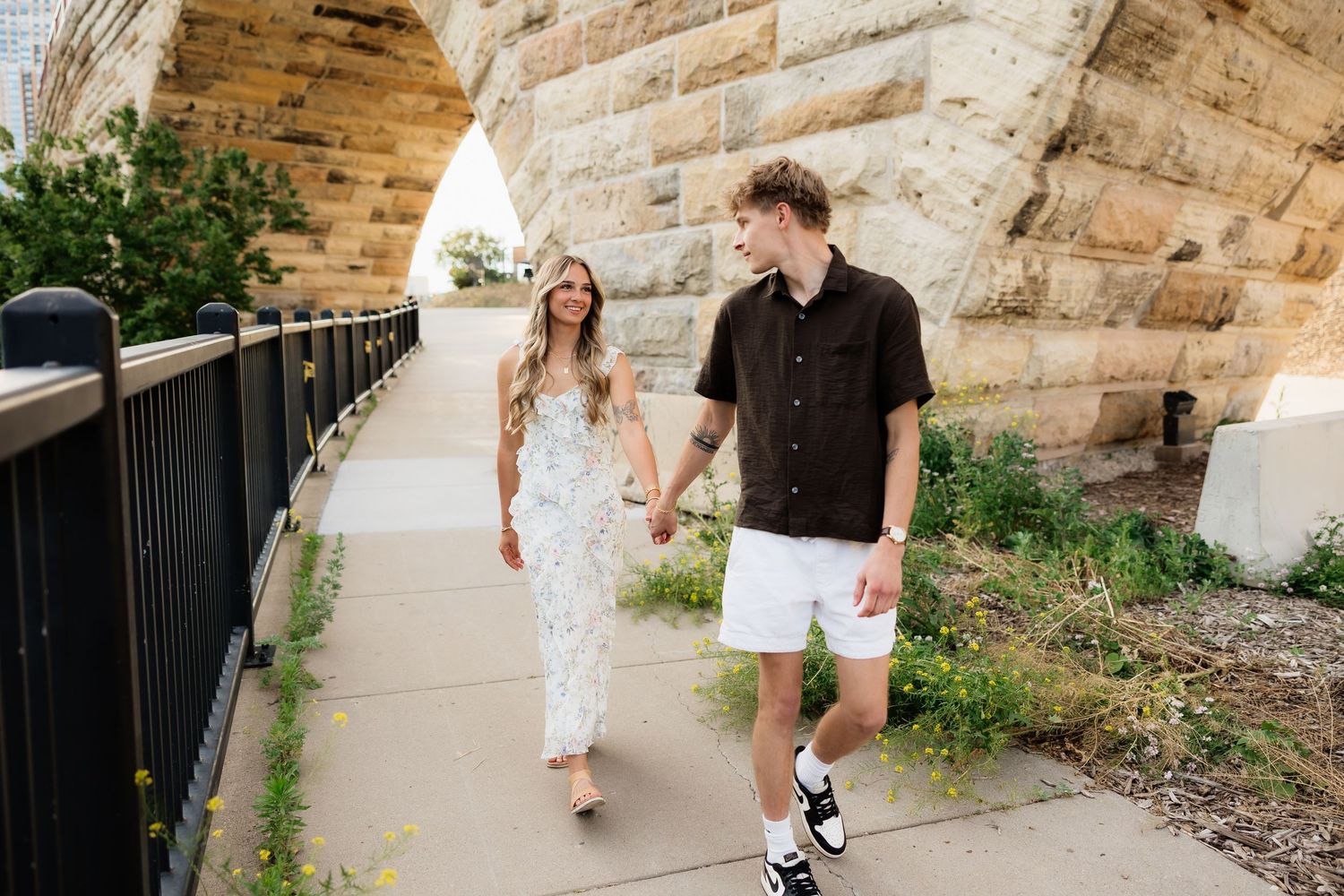 Couple holding hands walking on a sidewalk path near a stone arch bridge outdoors.