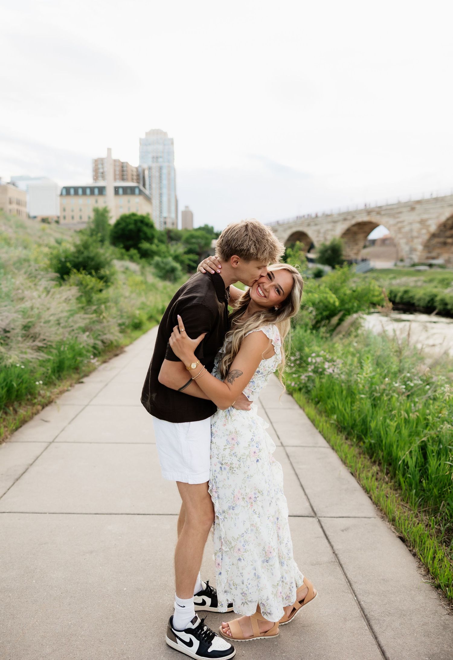 Couple sharing a sweet kiss on a riverside path with a stone arch bridge in the background.