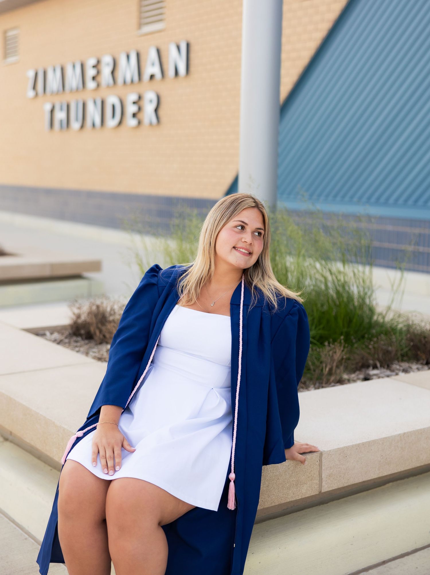 High school graduate in a blue gown sitting outside Zimmerman Thunder school building.