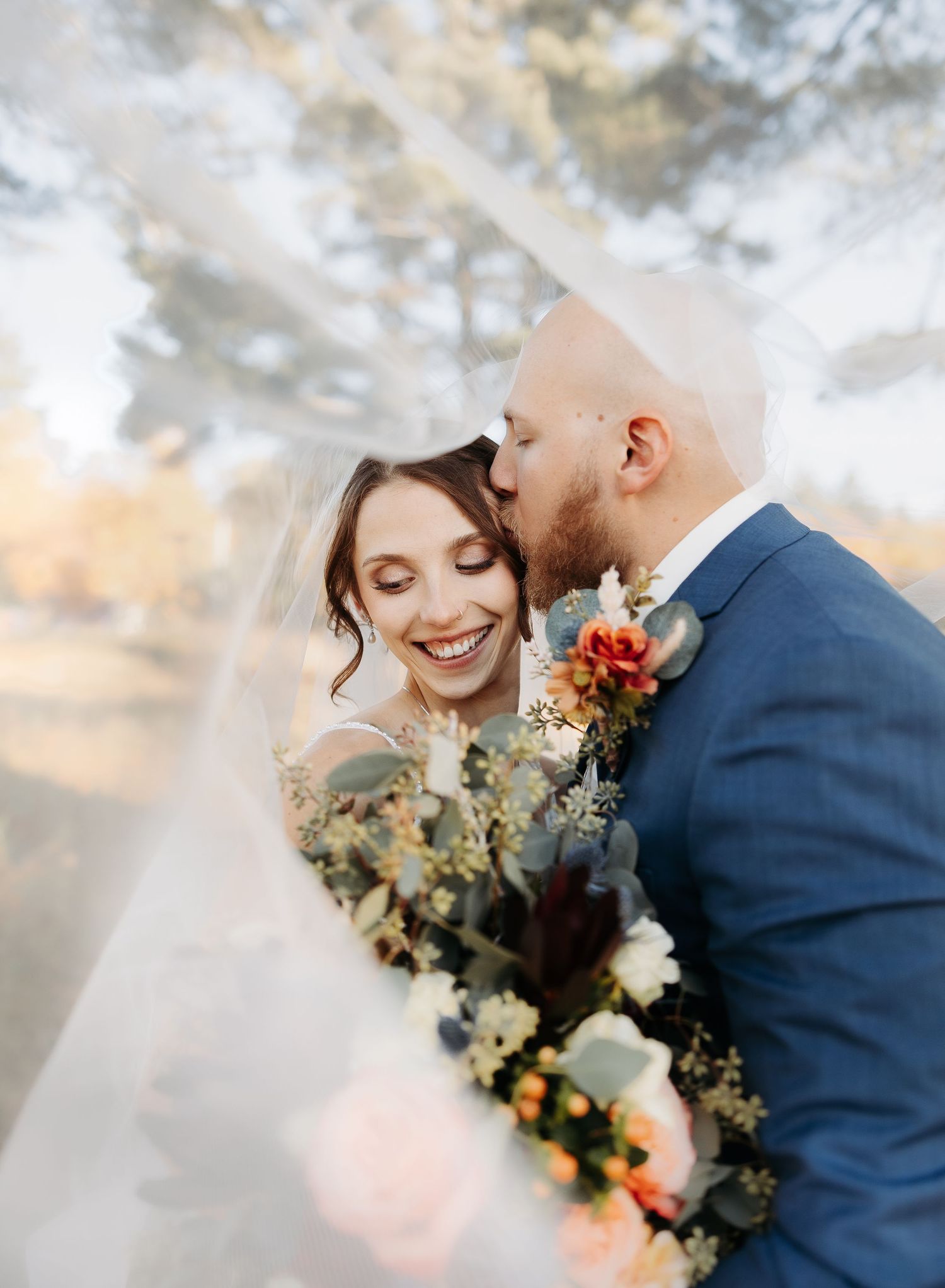 Groom in blue suit kissing smiling bride on cheek under flowing veil with colorful fall floral bouquet.