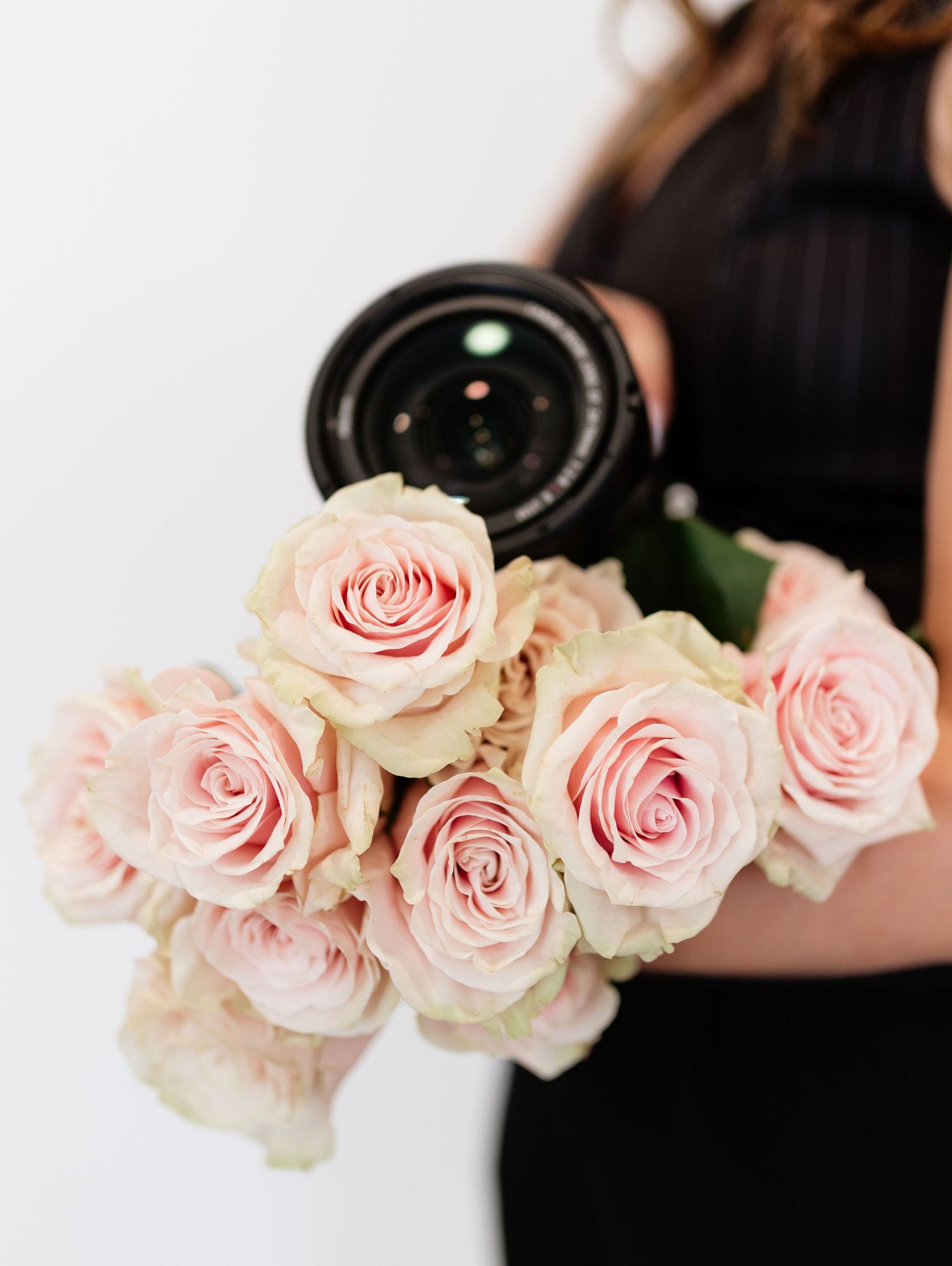Woman in black dress holding a camera and bouquet of soft pink roses against a white background.